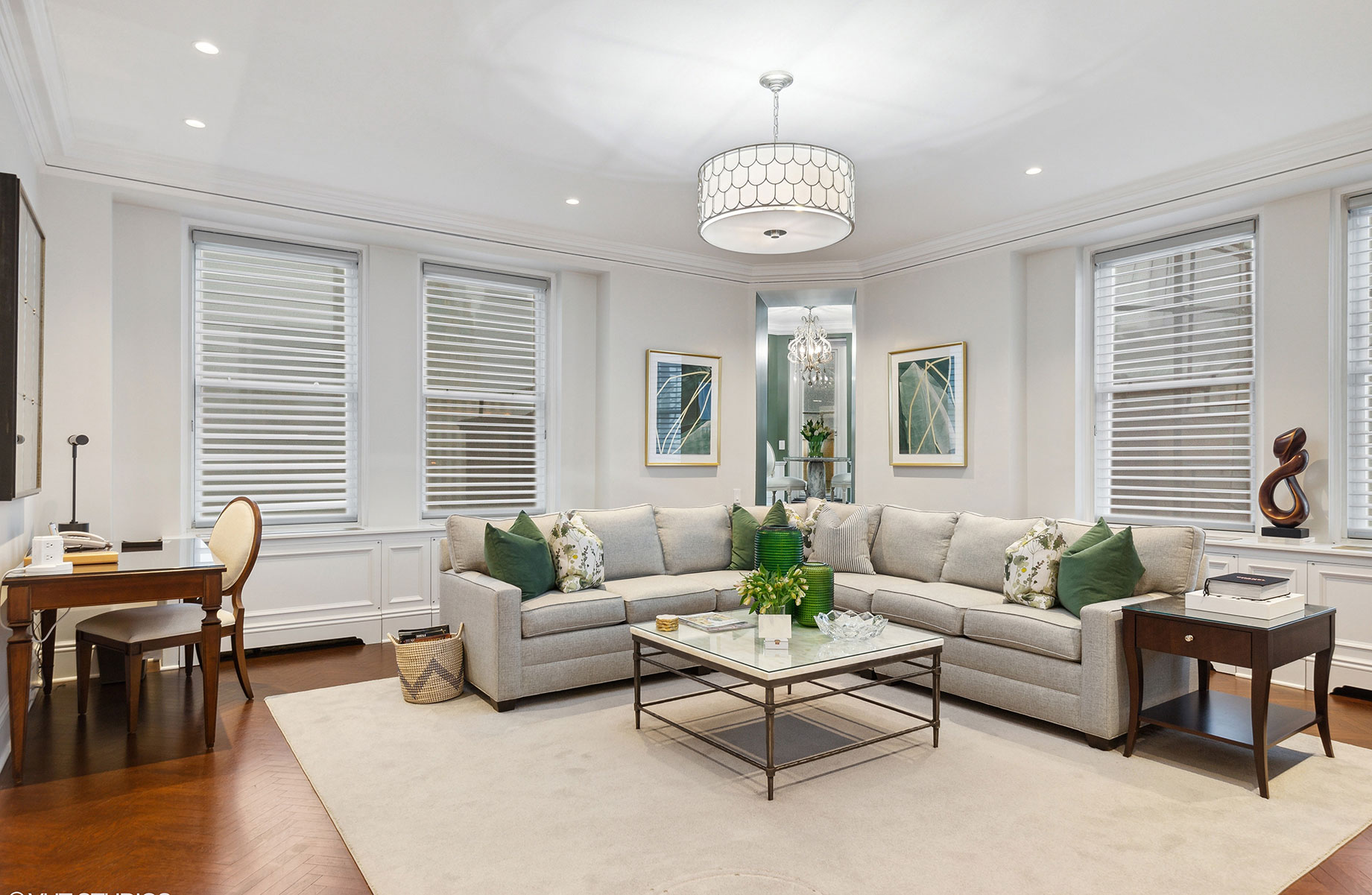 Contemporary living room with a neutral sectional sofa, green accent pillows, glass-top coffee table, wall art, and a decorative ceiling light fixture.