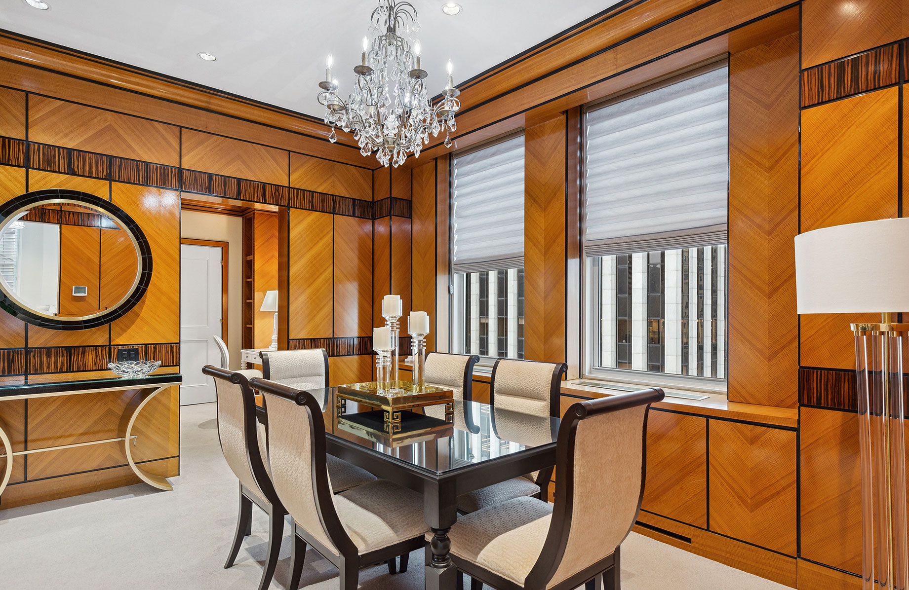 Luxurious dining area with a glass-top table and six upholstered chairs, surrounded by polished wood-paneled walls, modern window shades, and a crystal chandelier overhead.