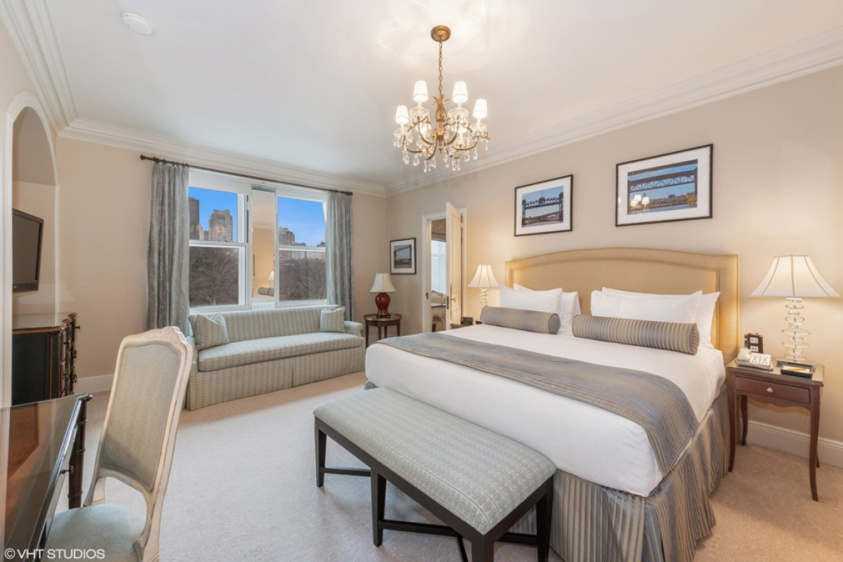Elegant hotel suite with a king-size bed, striped upholstered bench, and chandelier, featuring Central Park views through tall windows.
