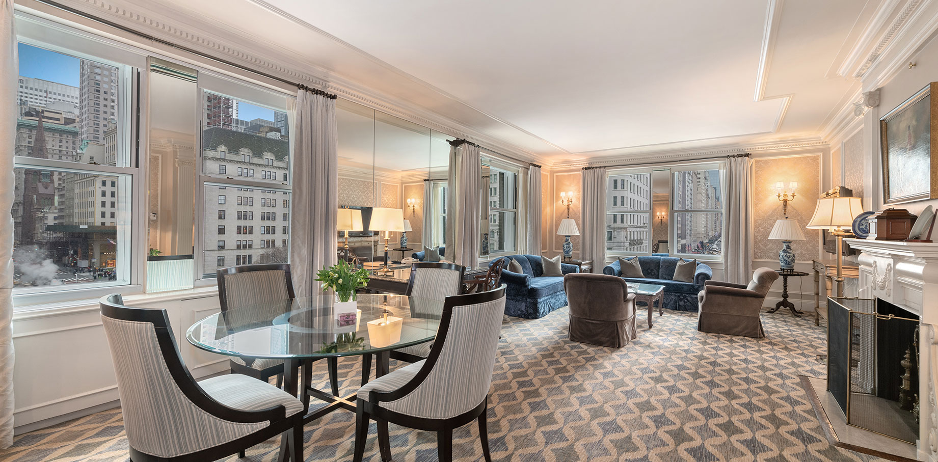 Spacious luxury hotel suite with patterned carpet, mirrored wall, round glass dining table, velvet seating, and panoramic views of a bustling city skyline through large windows.