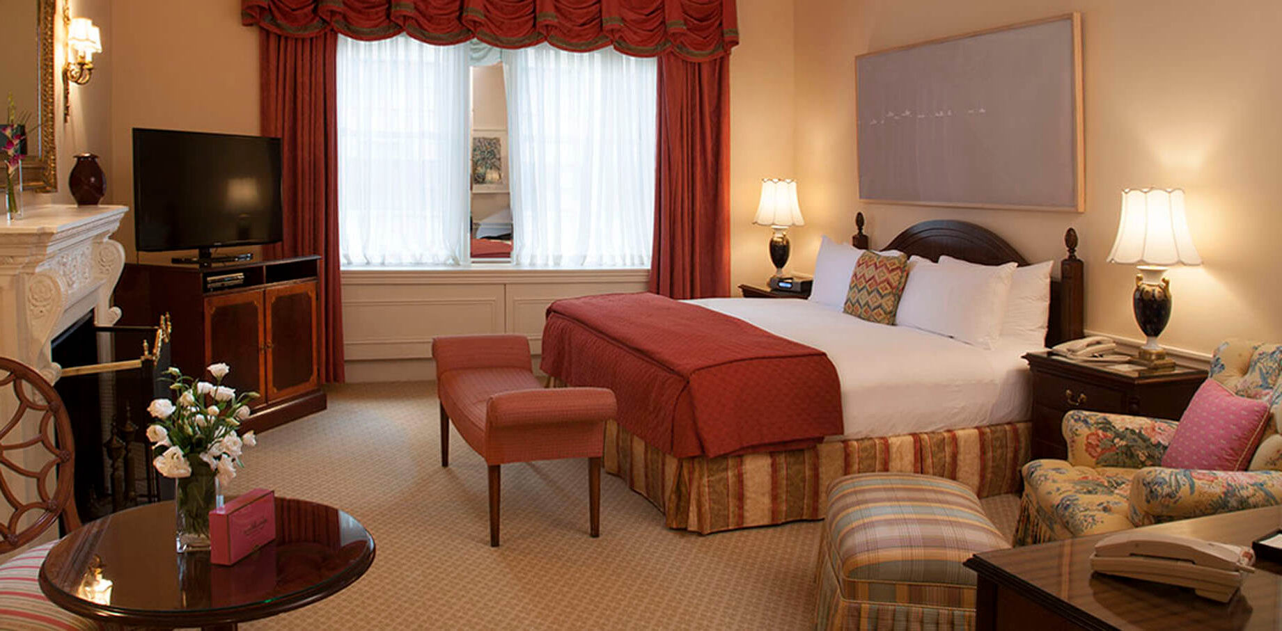 Warmly lit hotel room with a king bed, red bedding, floral armchair, striped bench, fireplace, flat-screen TV, and a large window draped with red curtains and sheer white panels.