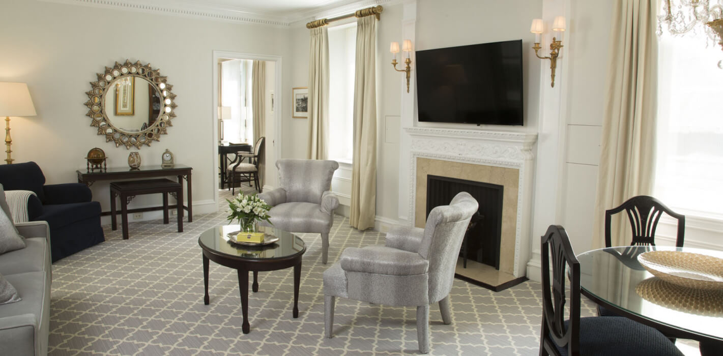Elegant living and dining area featuring a decorative fireplace with a flat-screen TV above, silver upholstered armchairs, a glass-top coffee table with fresh flowers, and a dark wood dining set under a crystal chandelier.