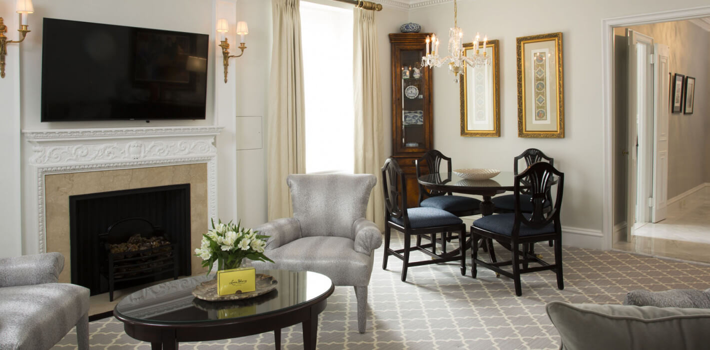 Elegant living and dining area featuring a decorative fireplace with a flat-screen TV above, silver upholstered armchairs, a glass-top coffee table with fresh flowers, and a dark wood dining set under a crystal chandelier.