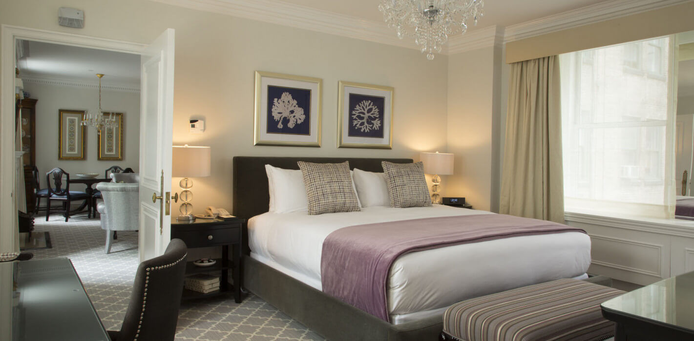 Sophisticated hotel bedroom with a plush king bed adorned in white linens and a lavender throw, flanked by crystal lamps and modern art, with an adjoining dining area visible through an open door.