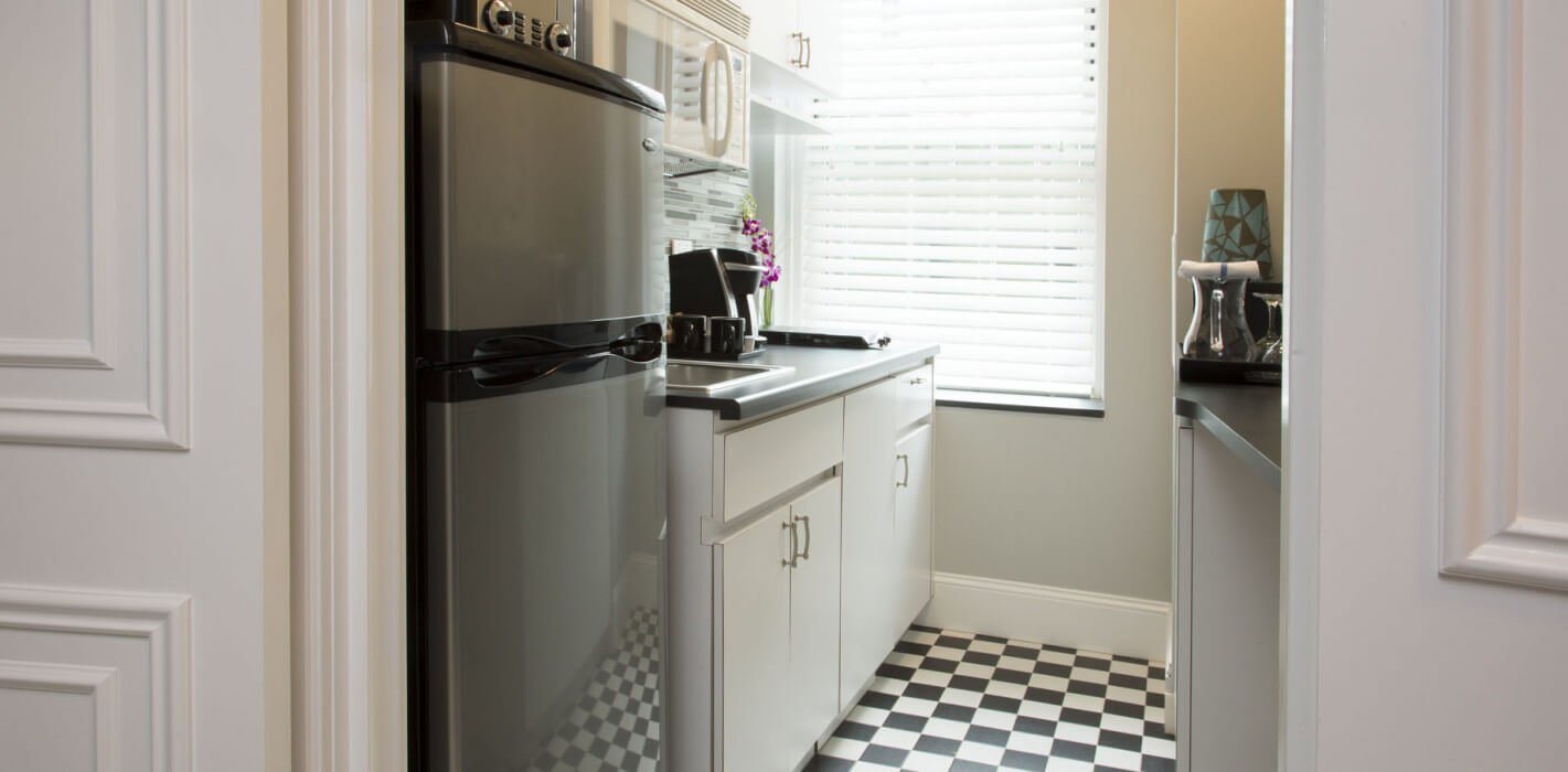 Compact galley kitchen featuring black-and-white checkered flooring, stainless steel appliances, white cabinetry, and a bright window with horizontal blinds.