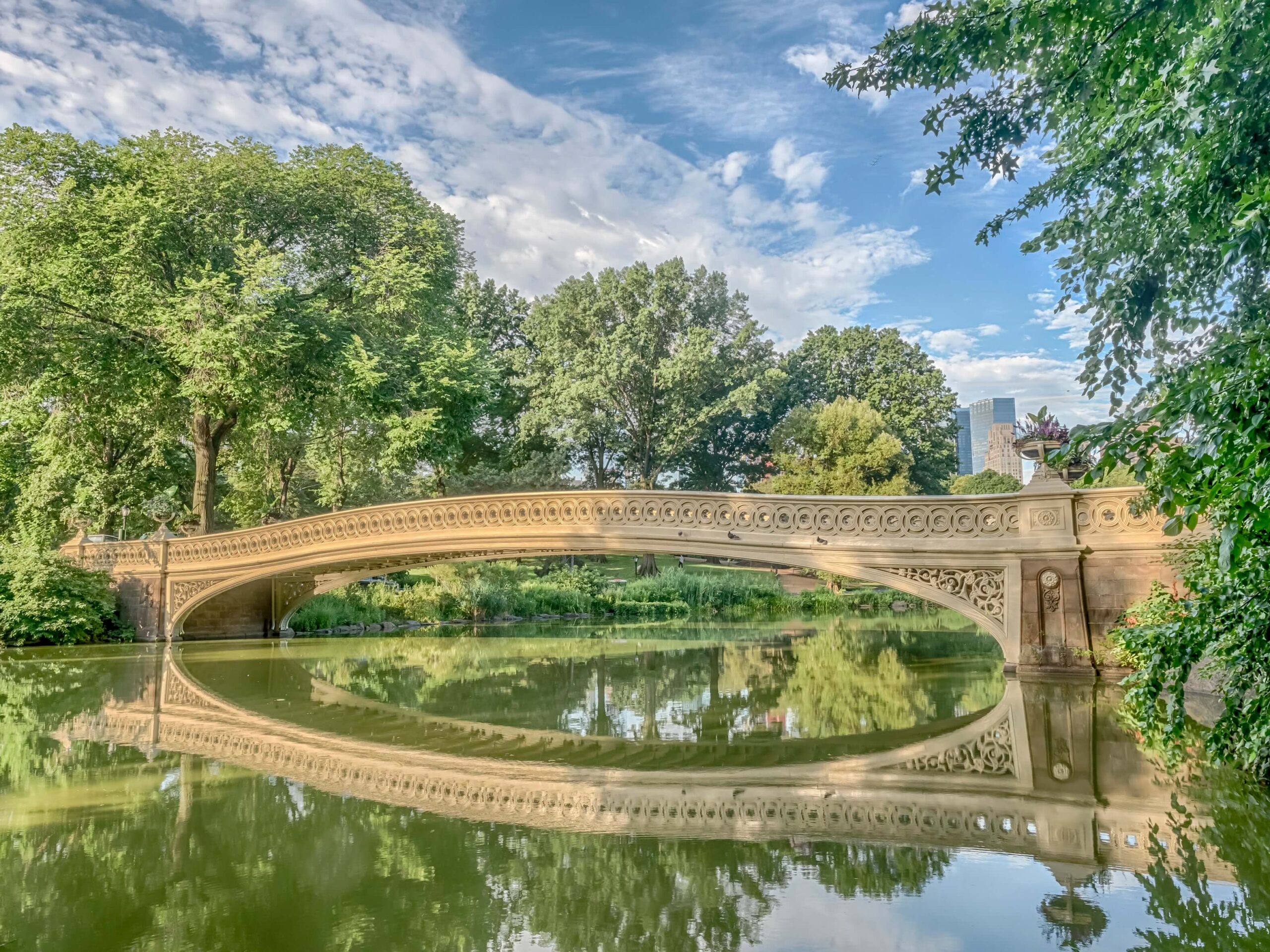 Bow Bridge in Central Park reflecting on the lake, surrounded by greenery and New York City skyline