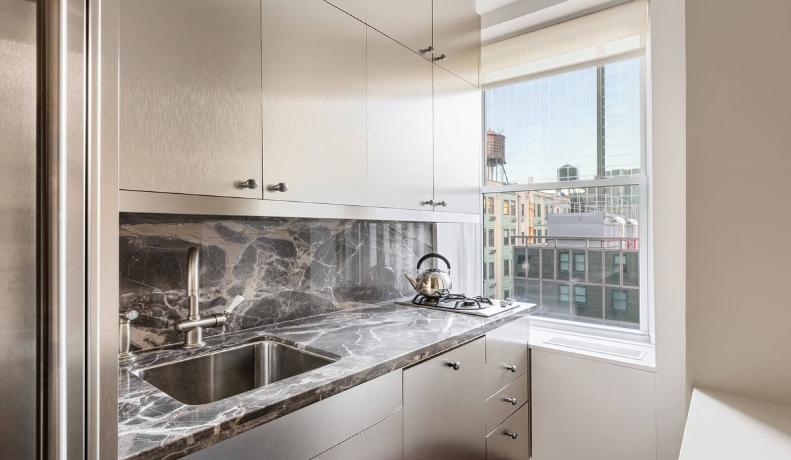 Modern galley kitchen with sleek stainless steel cabinetry, a dark marble countertop and backsplash, built-in gas cooktop, and a window overlooking Midtown Manhattan.