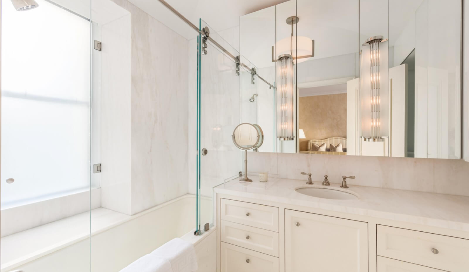 Bright, modern bathroom with a marble vanity, mirrored cabinets, a built-in bathtub with glass sliding doors, and soft overhead lighting.