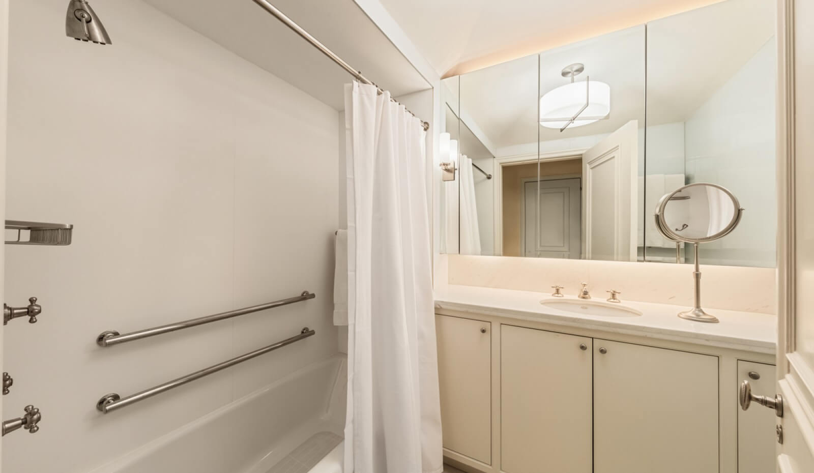 Elegant bathroom featuring a white shower-tub combo with safety grab bars, a curtain, a wide vanity with under-sink cabinets, and mirrored walls with modern lighting.