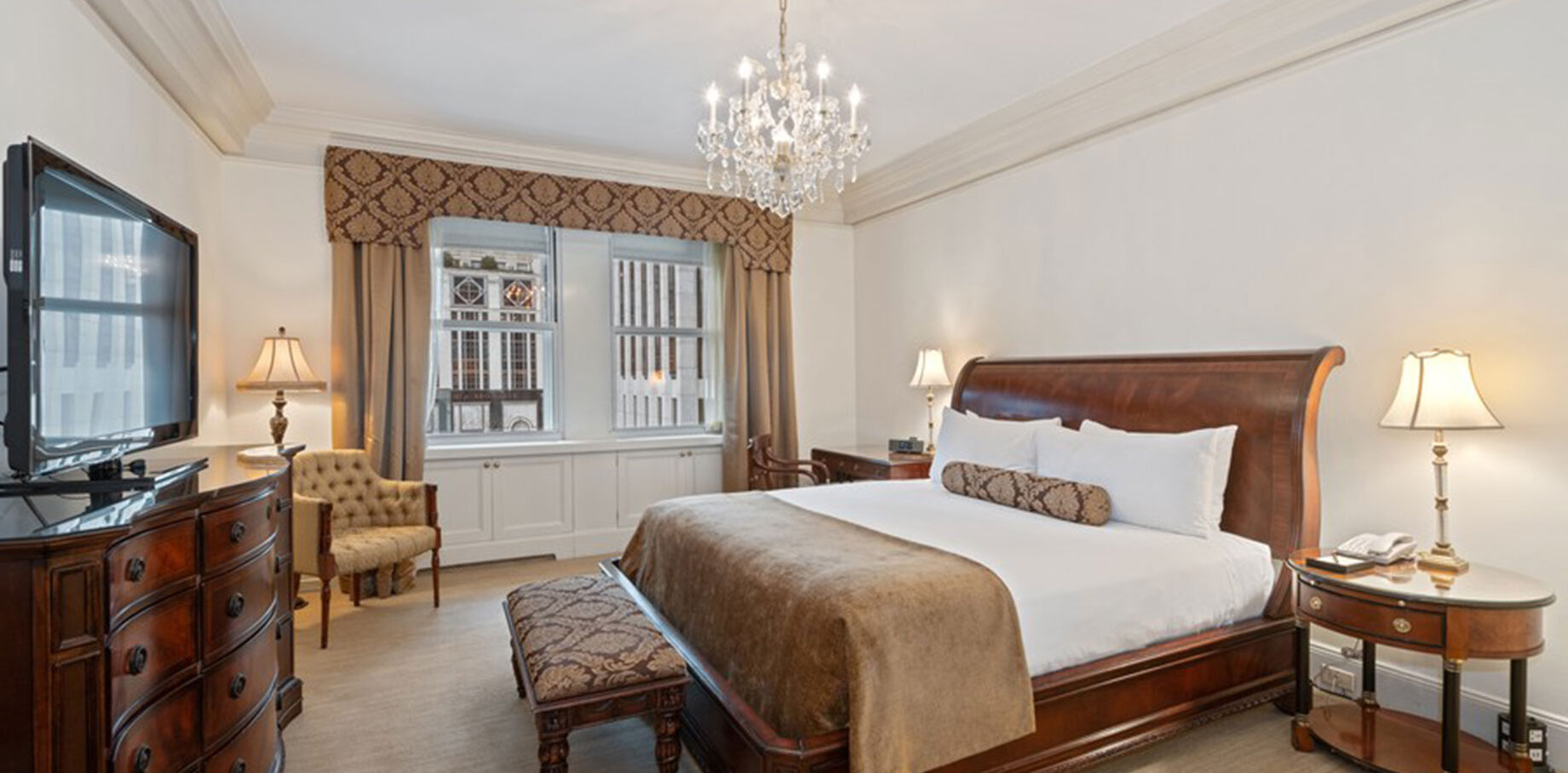 Refined hotel bedroom with a sleigh-style king bed, crystal chandelier, classic wood furnishings, and windows facing urban architecture.