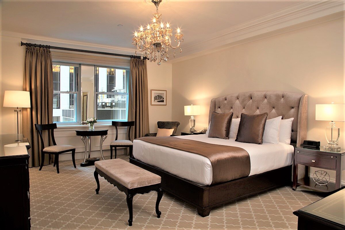 Elegant hotel room featuring a tufted headboard king bed with satin bedding, crystal chandelier, dark wood furniture, and a sitting area near a city-facing window.