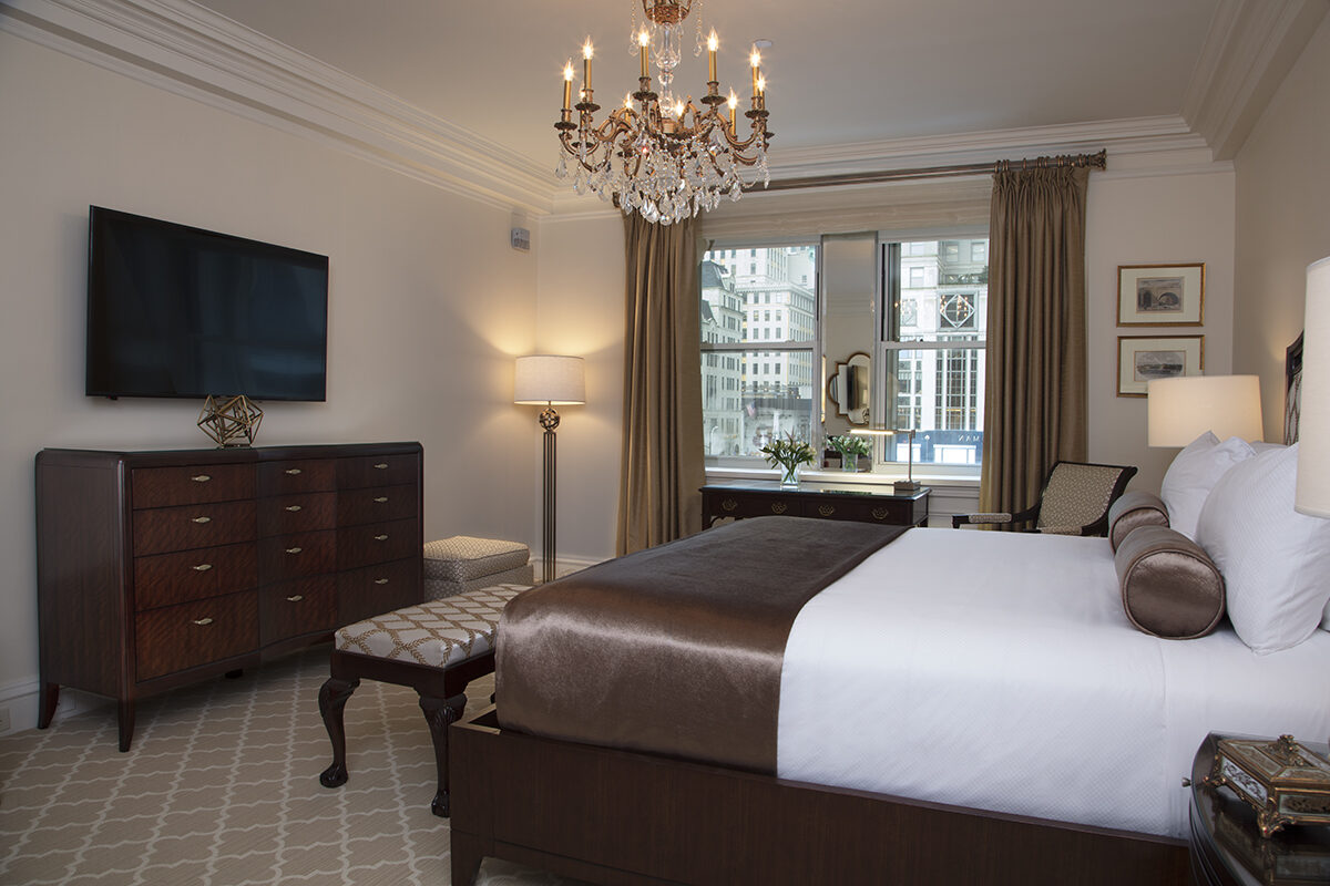 Sophisticated hotel room with a plush king bed, chocolate satin bedding, crystal chandelier, and city view through a large window with taupe curtains.