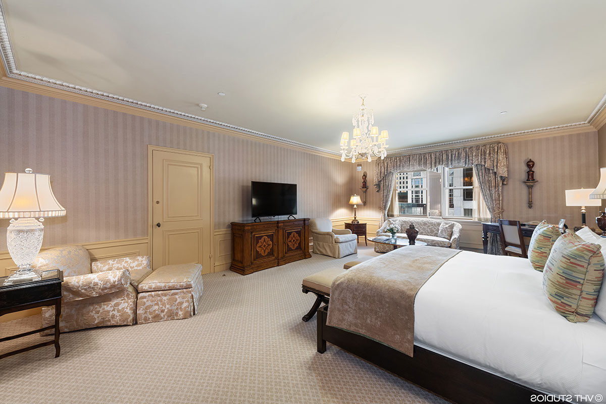 Spacious traditional hotel suite with a king bed, plush seating area, crystal chandelier, classic wood furnishings, and elegant patterned wallpaper.