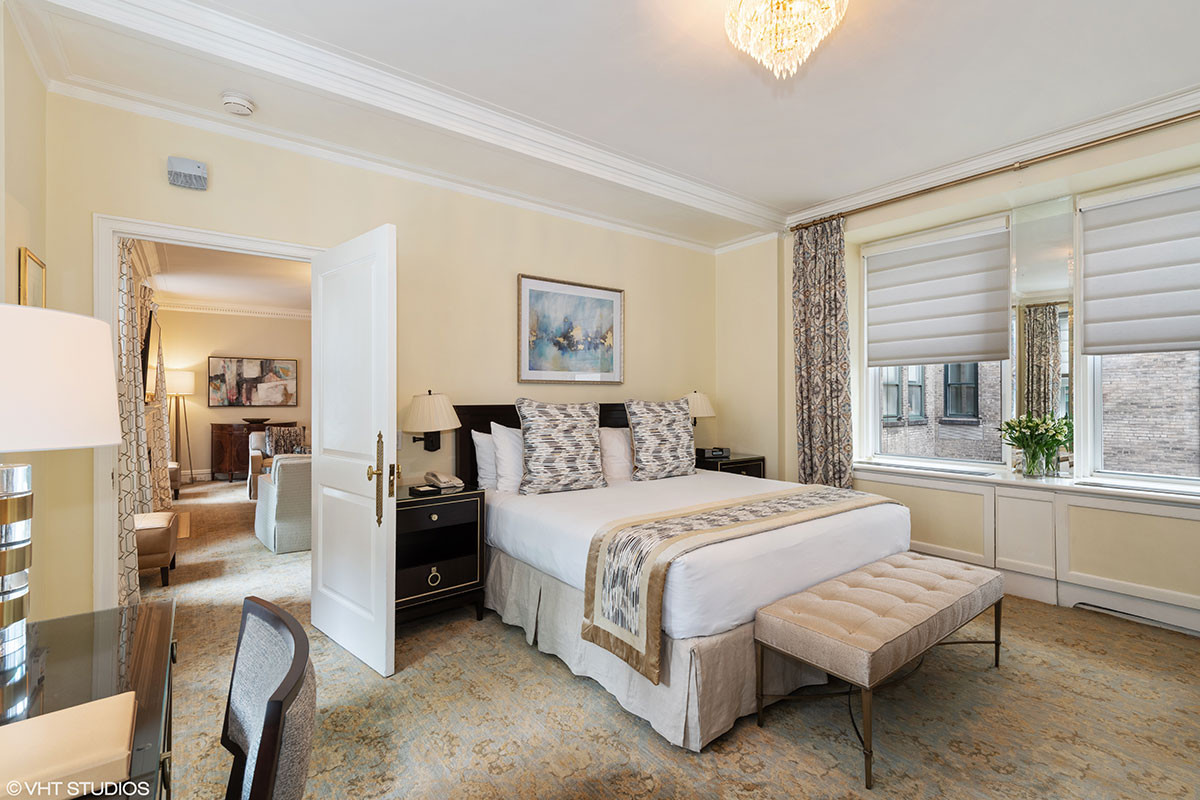 Bright and elegant hotel bedroom featuring a king-size bed with geometric accent pillows, a tufted bench, and large windows with Roman shades, connected to a separate living area through an open doorway.