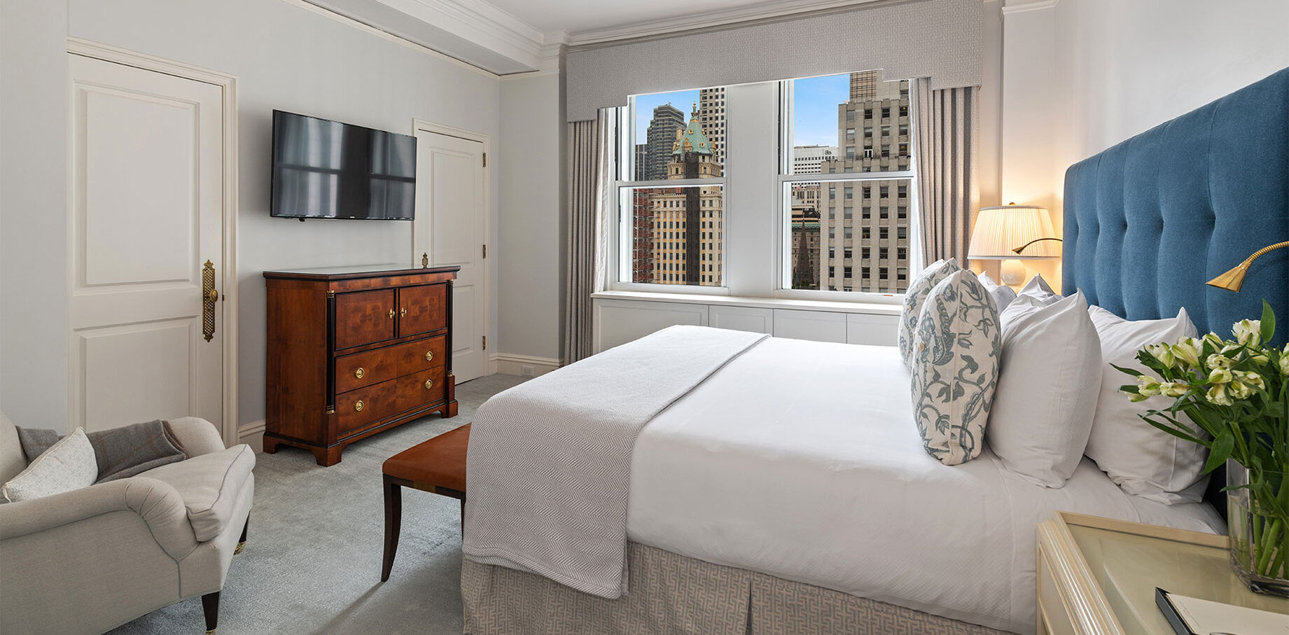 Bright hotel bedroom featuring a plush blue tufted headboard, elegant wood furniture, and large windows with views of Manhattan skyscrapers.