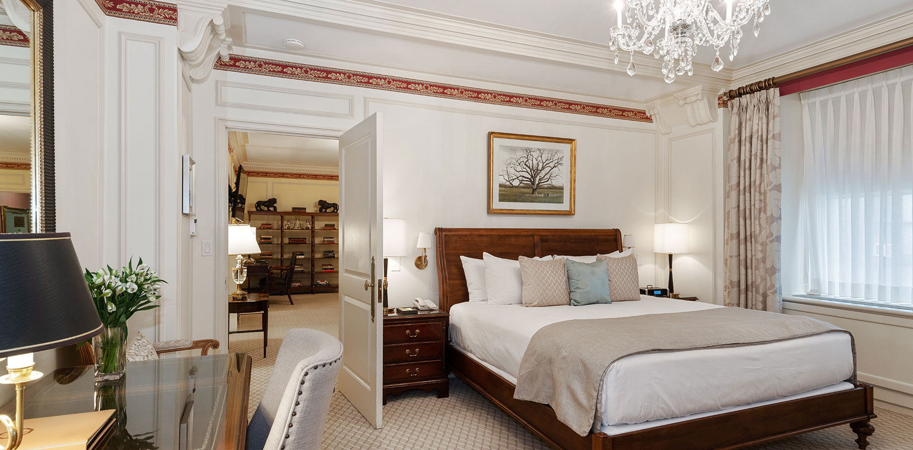 Elegant hotel bedroom with a wooden sleigh bed, crystal chandelier, writing desk, and partial view into a richly appointed sitting room beyond.