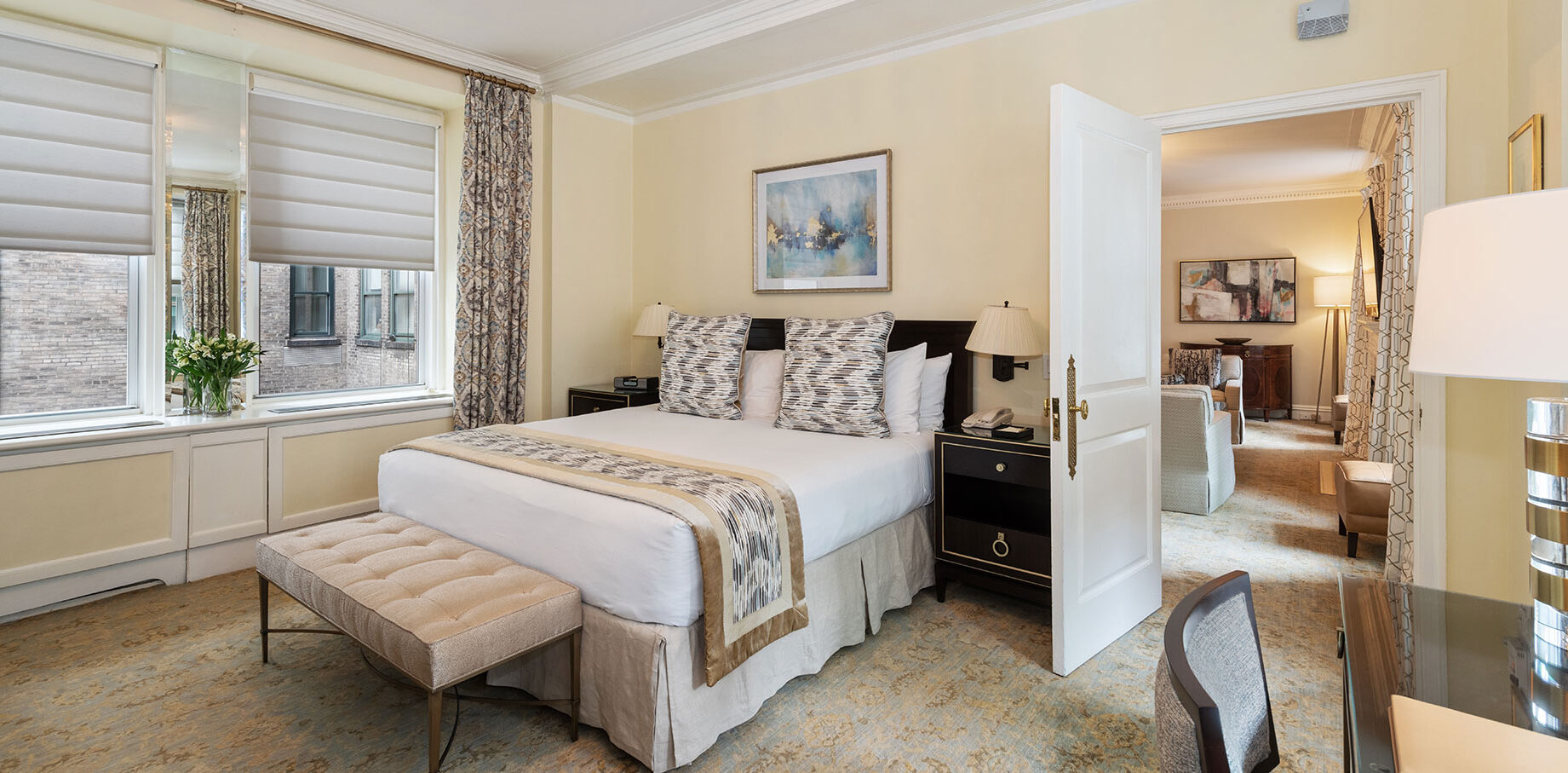 Charming hotel bedroom with patterned pillows, a tufted bench at the foot of the bed, light-filtering roman shades, and an open door revealing a contemporary suite beyond.