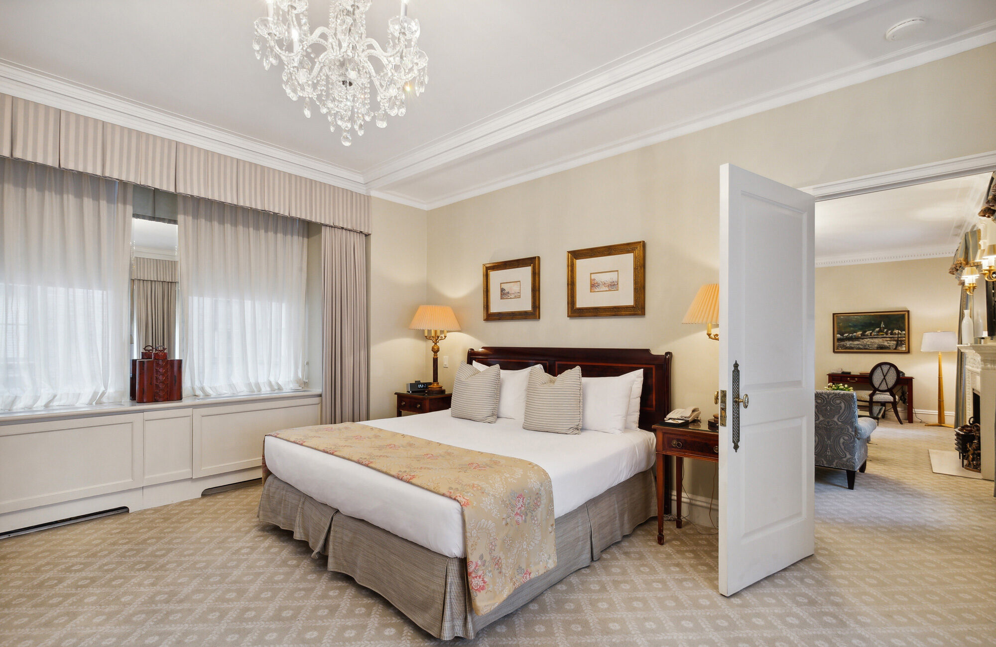 Elegant hotel bedroom with a classic wood-framed bed, light-patterned bed runner, crystal chandelier, and open door revealing a connected sitting room with traditional décor.