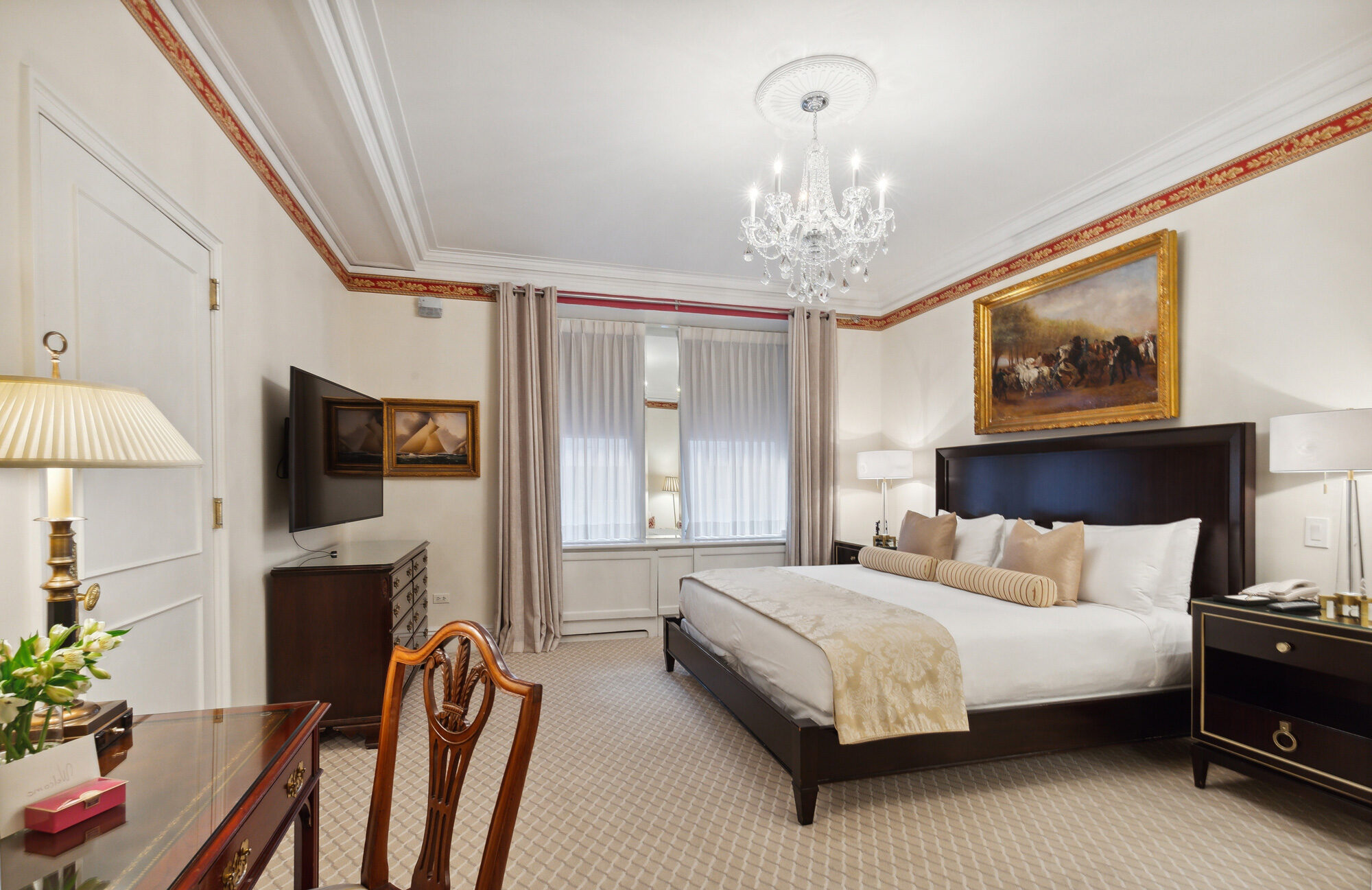Traditional hotel room with a crystal chandelier, king bed, dark wood furnishings, gold-framed classical artwork, and a decorative border running along the ceiling.
