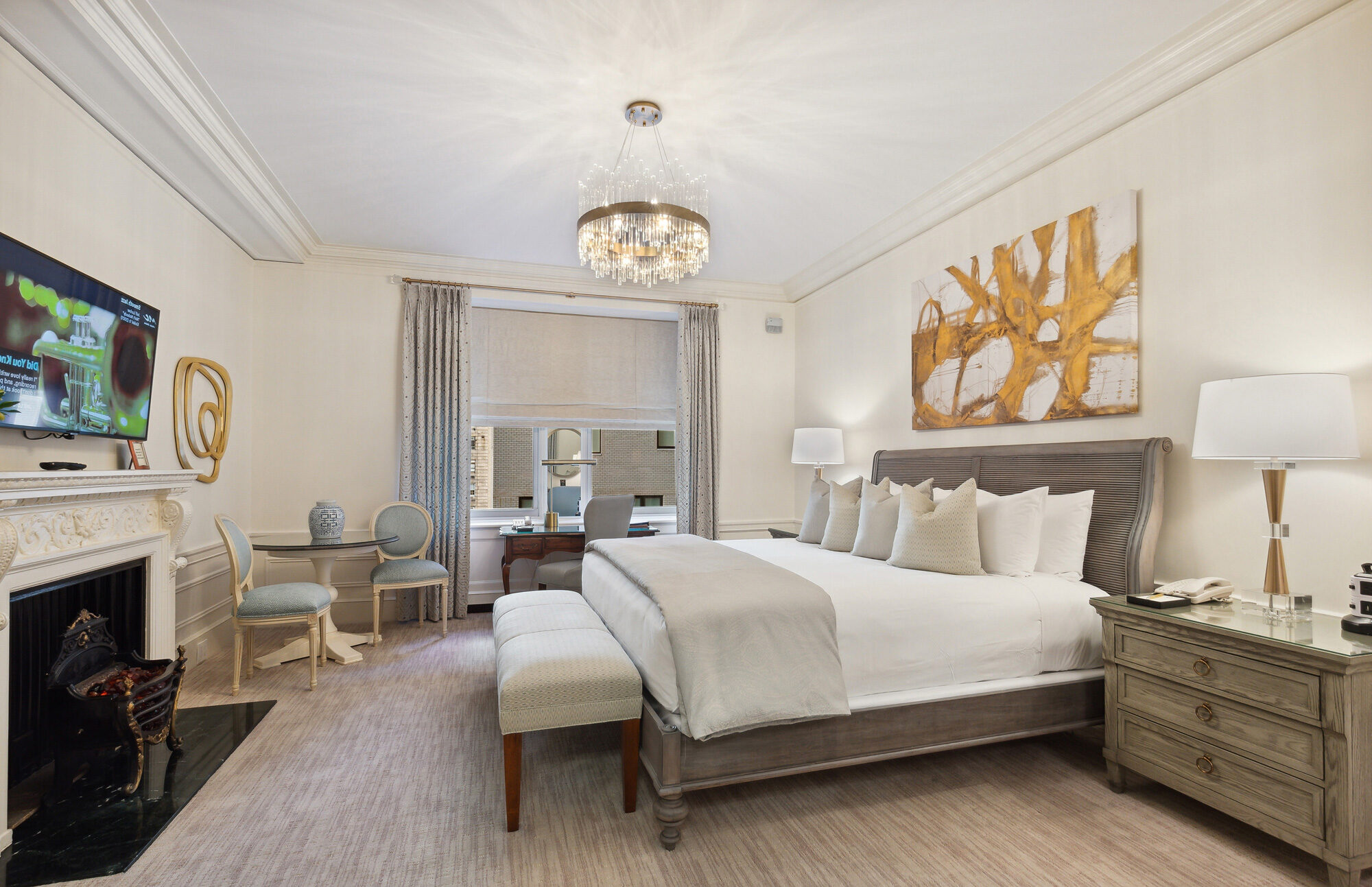 Bright, contemporary hotel room featuring a king bed with neutral-toned linens, modern abstract artwork, crystal chandelier, fireplace, and sitting area with classic chairs.