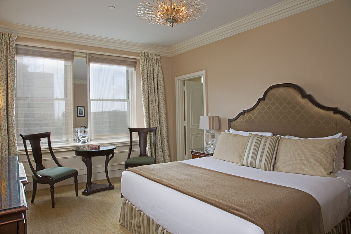 Bright guest room featuring a king bed with a diamond-tufted headboard, neutral-toned linens, two wood chairs and a table near tall windows with natural light.