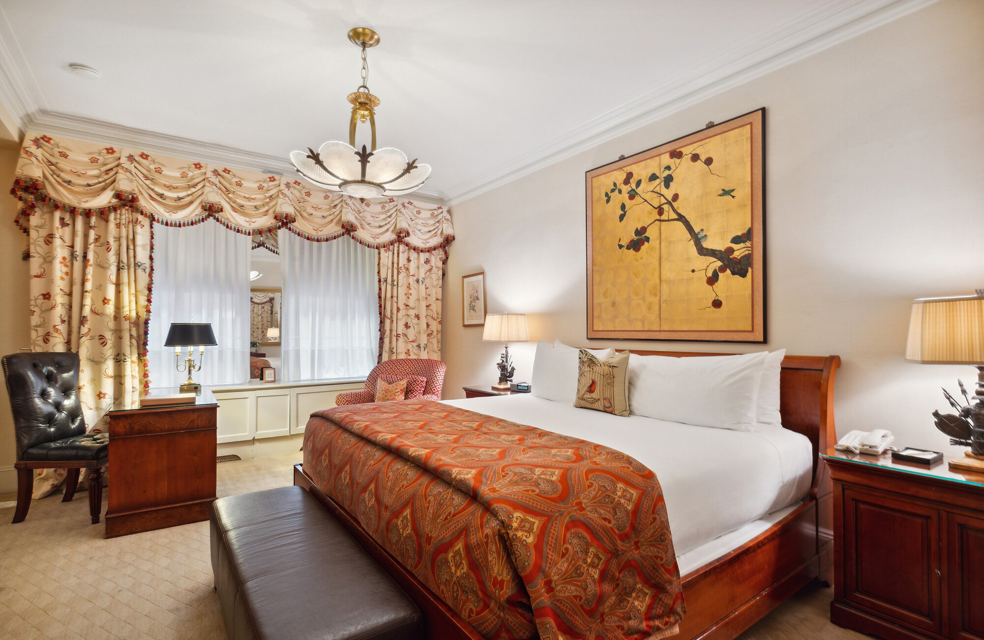 Elegant hotel room featuring a sleigh bed with a patterned red coverlet, a large Japanese-style botanical wall painting, ornate floral curtains, and vintage furnishings with warm wood finishes.