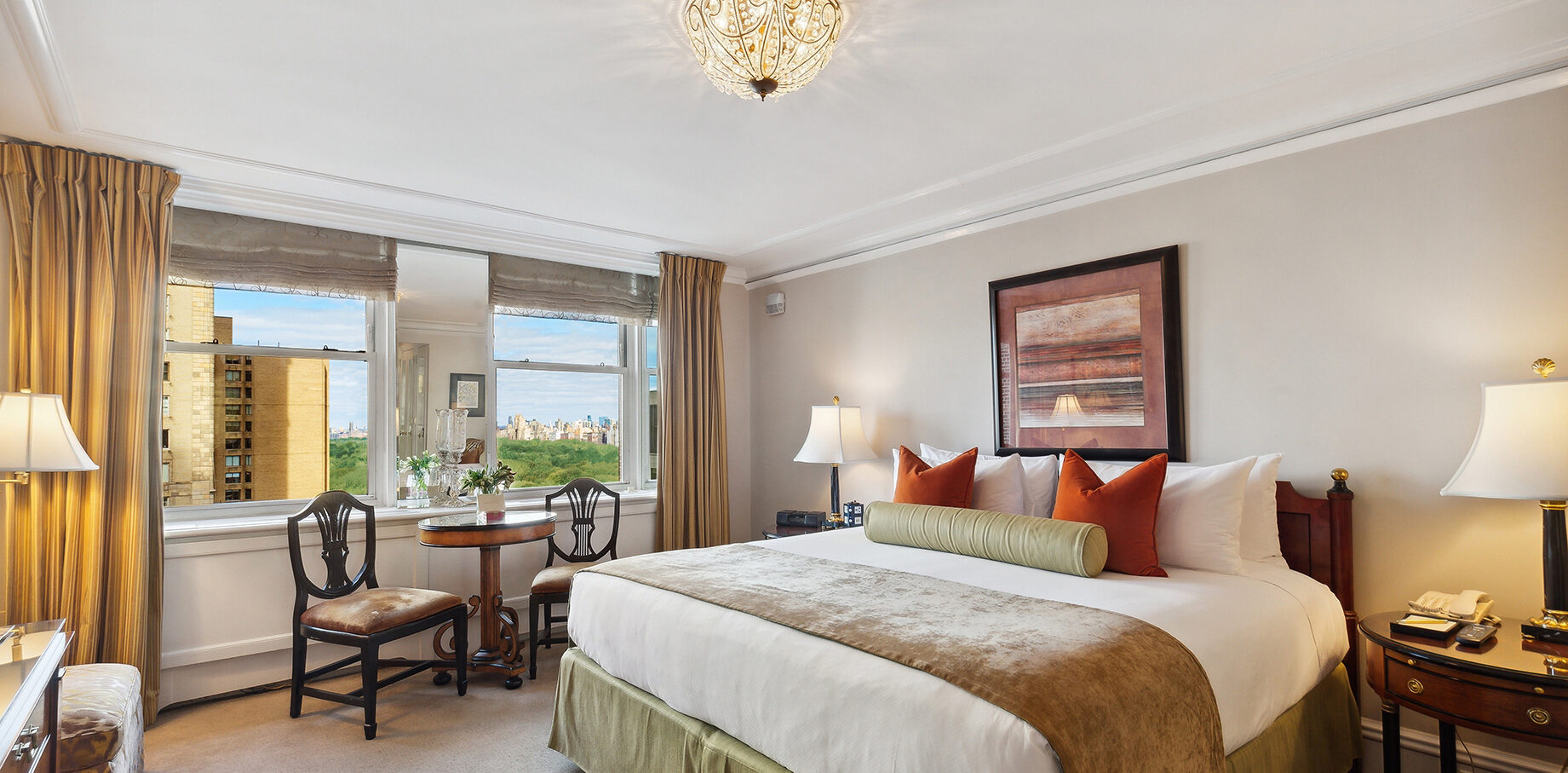 Sunlit hotel room with a king bed, bold accent pillows, classic wood furnishings, and panoramic windows overlooking Central Park and the city skyline.
