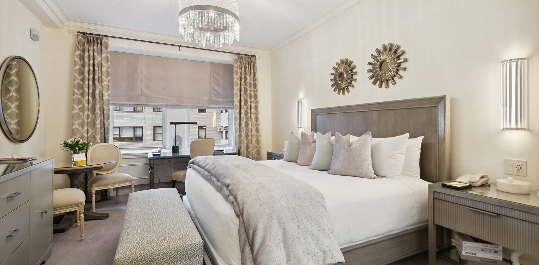 Bright hotel room with soft neutral tones, plush bedding, metallic sunburst wall décor, and a crystal chandelier, featuring a seating area and large window with patterned drapes.