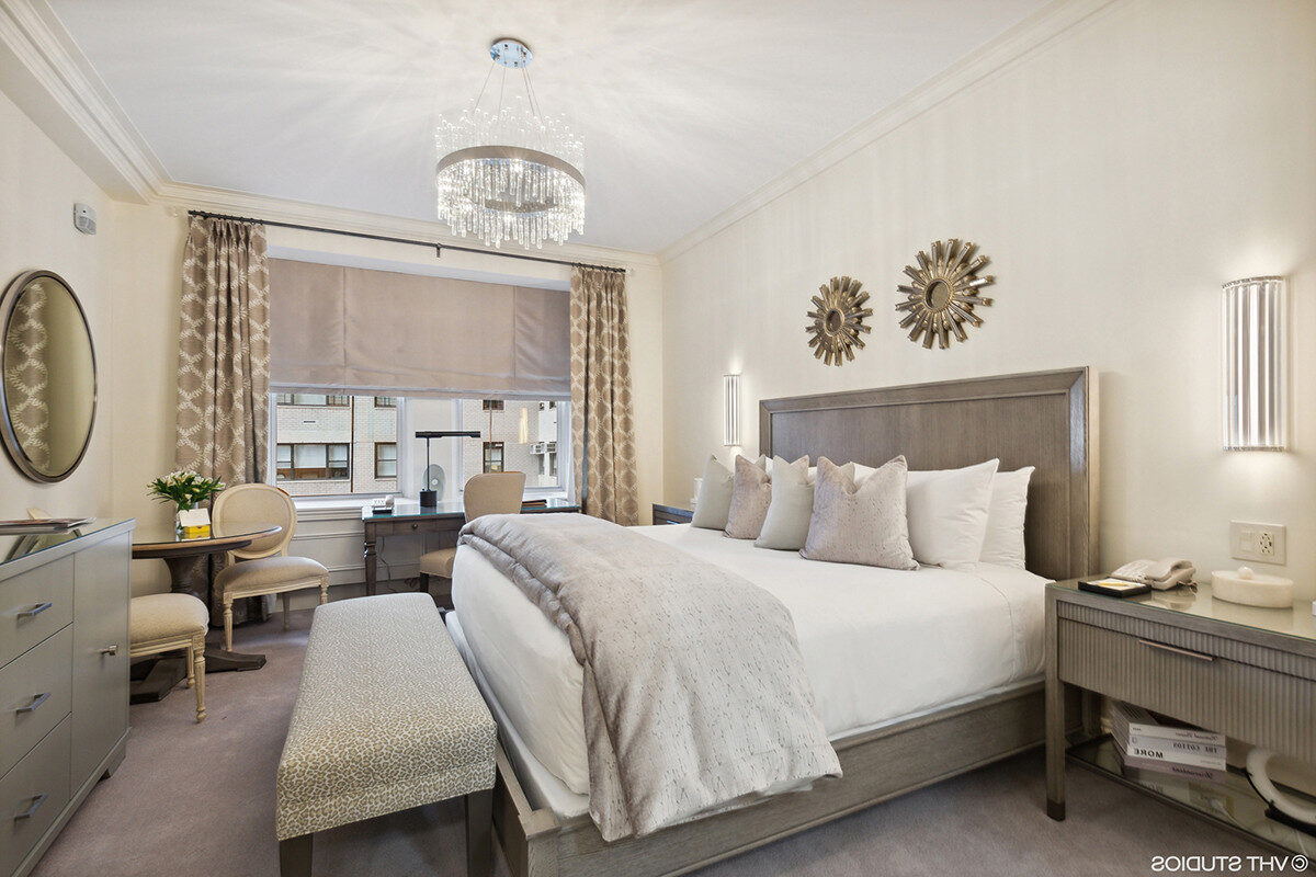 Contemporary hotel bedroom featuring a king bed with light gray linens, plush pillows, sunburst wall art, a crystal chandelier, and soft neutral-toned furnishings.