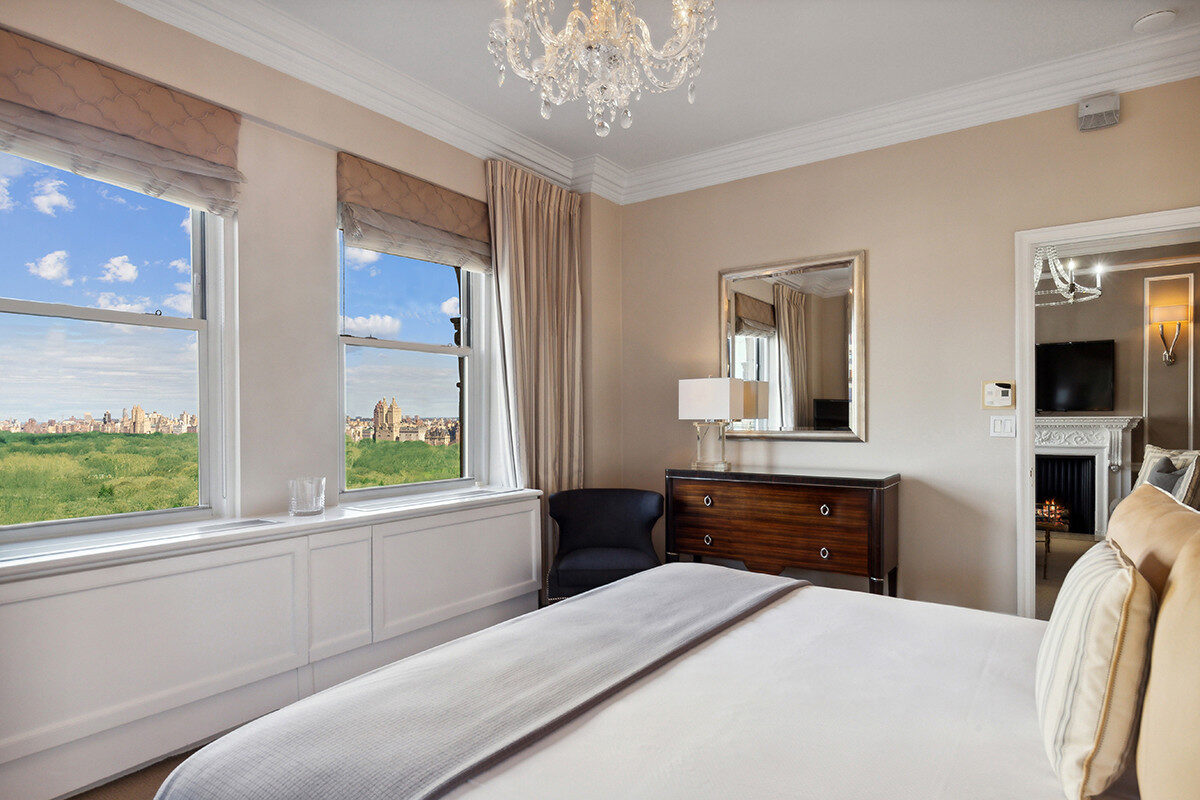 Luxurious hotel bedroom with beige walls and elegant crown molding, featuring a large bed with neutral linens, a crystal chandelier, a dark wood dresser with a mirror, and expansive windows offering sweeping views of Central Park and the Manhattan skyline.
