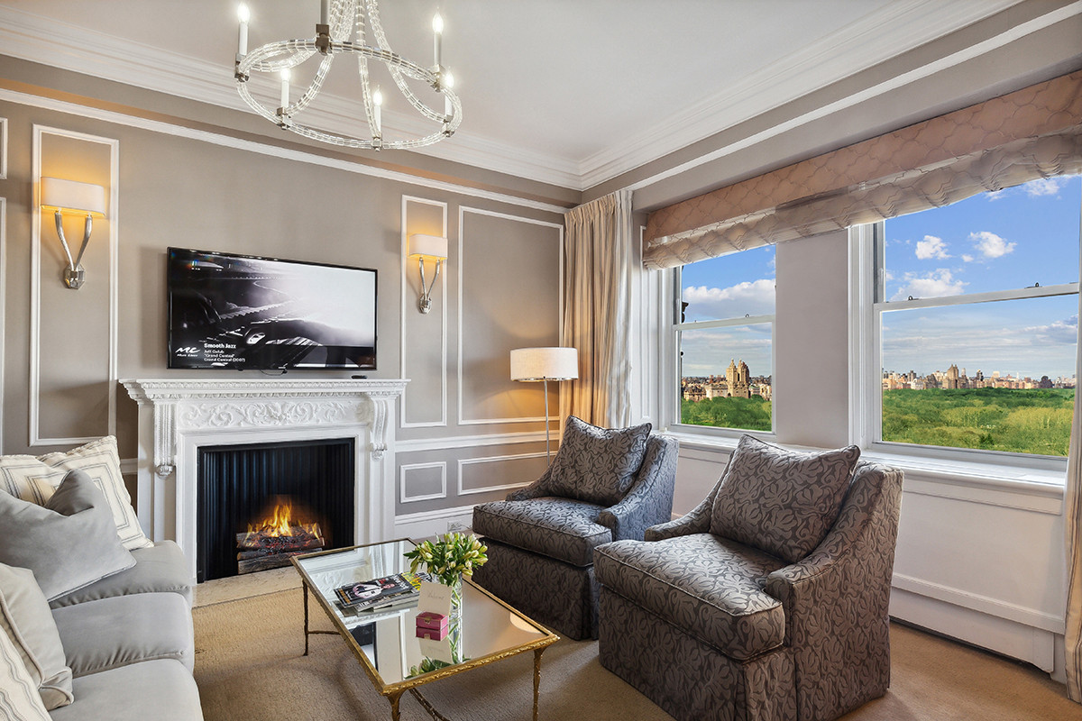 Elegant hotel suite with a gray-toned interior, decorative wall molding, plush armchairs, a white fireplace with a lit fire, wall-mounted TV, and large windows offering panoramic views of Central Park and the New York City skyline.