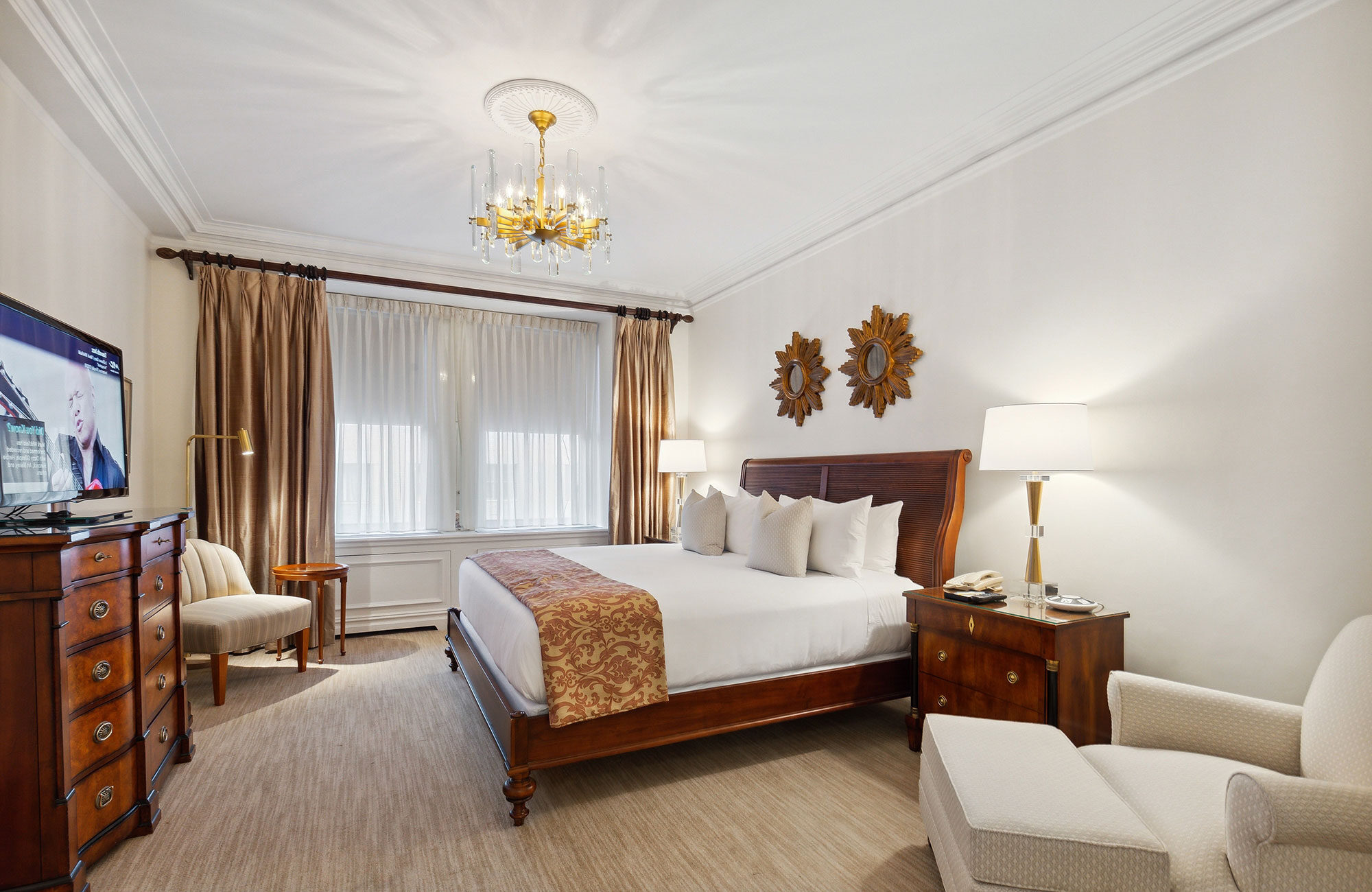Classic hotel bedroom featuring a king bed with wood frame, neutral linens and pillows, traditional chandelier, sunburst wall decor, and large curtained windows.