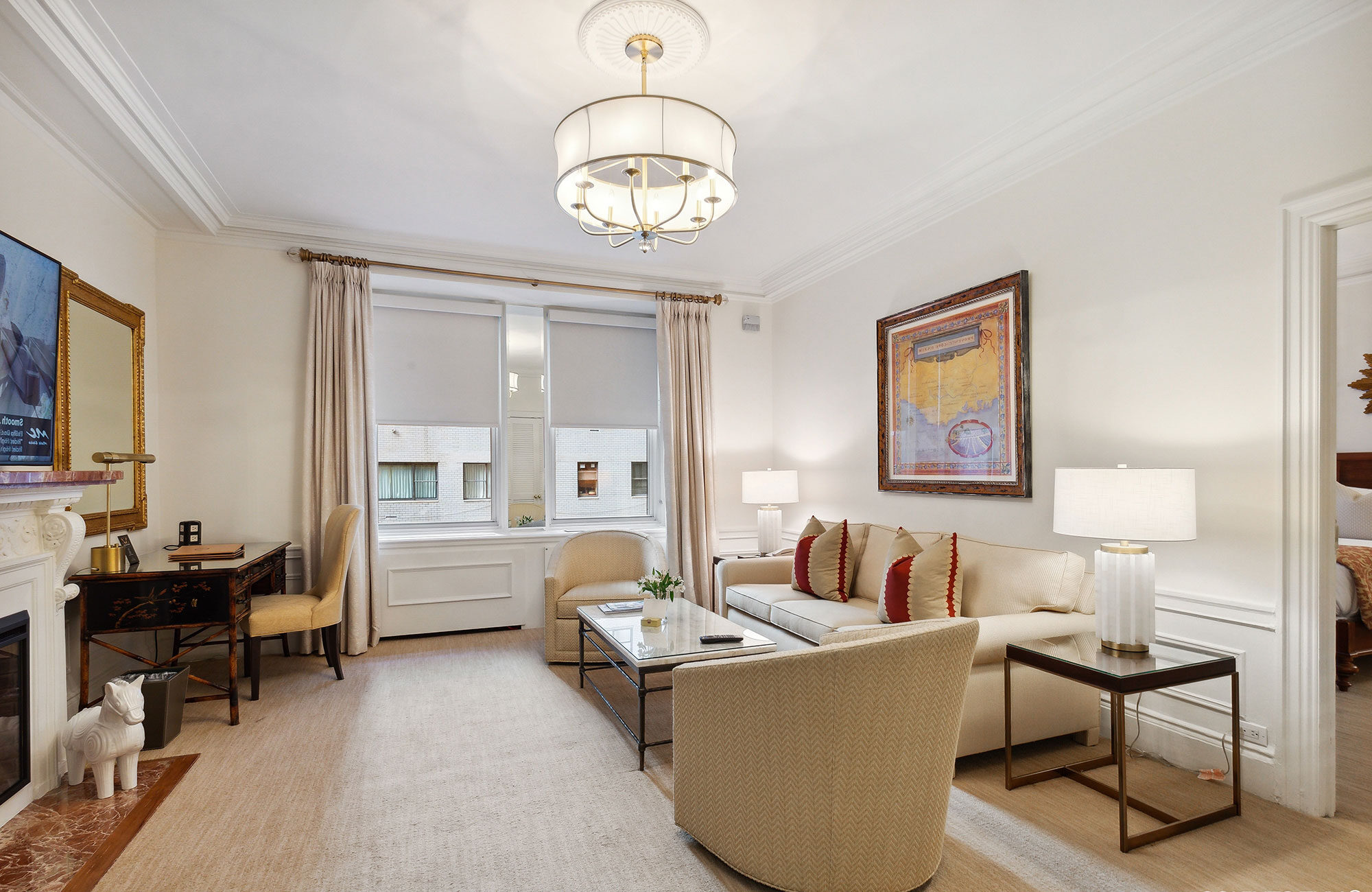 Refined hotel living room with a cream-colored sofa, modern chandelier, fireplace, classic writing desk, and large windows with natural light and neutral curtains.