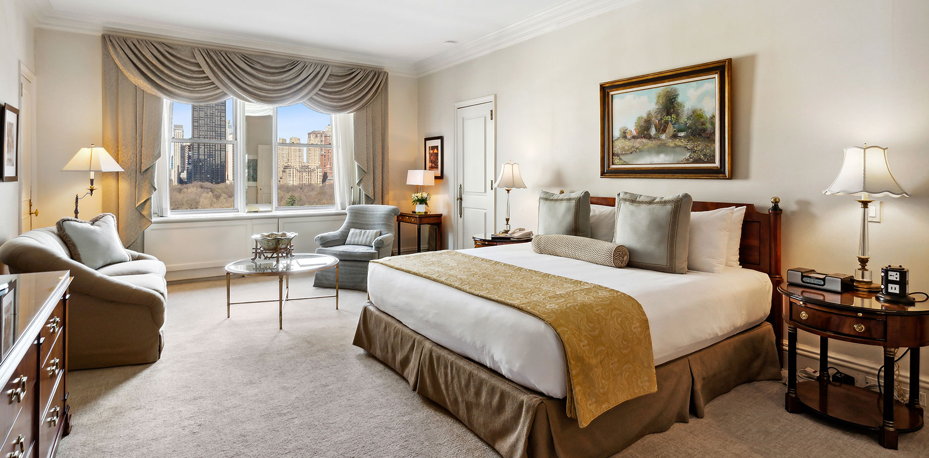 Elegant hotel room featuring a king bed with golden accents, classic furnishings, a seating area with armchairs and a glass-top coffee table, and a large window overlooking Central Park and Manhattan skyline.