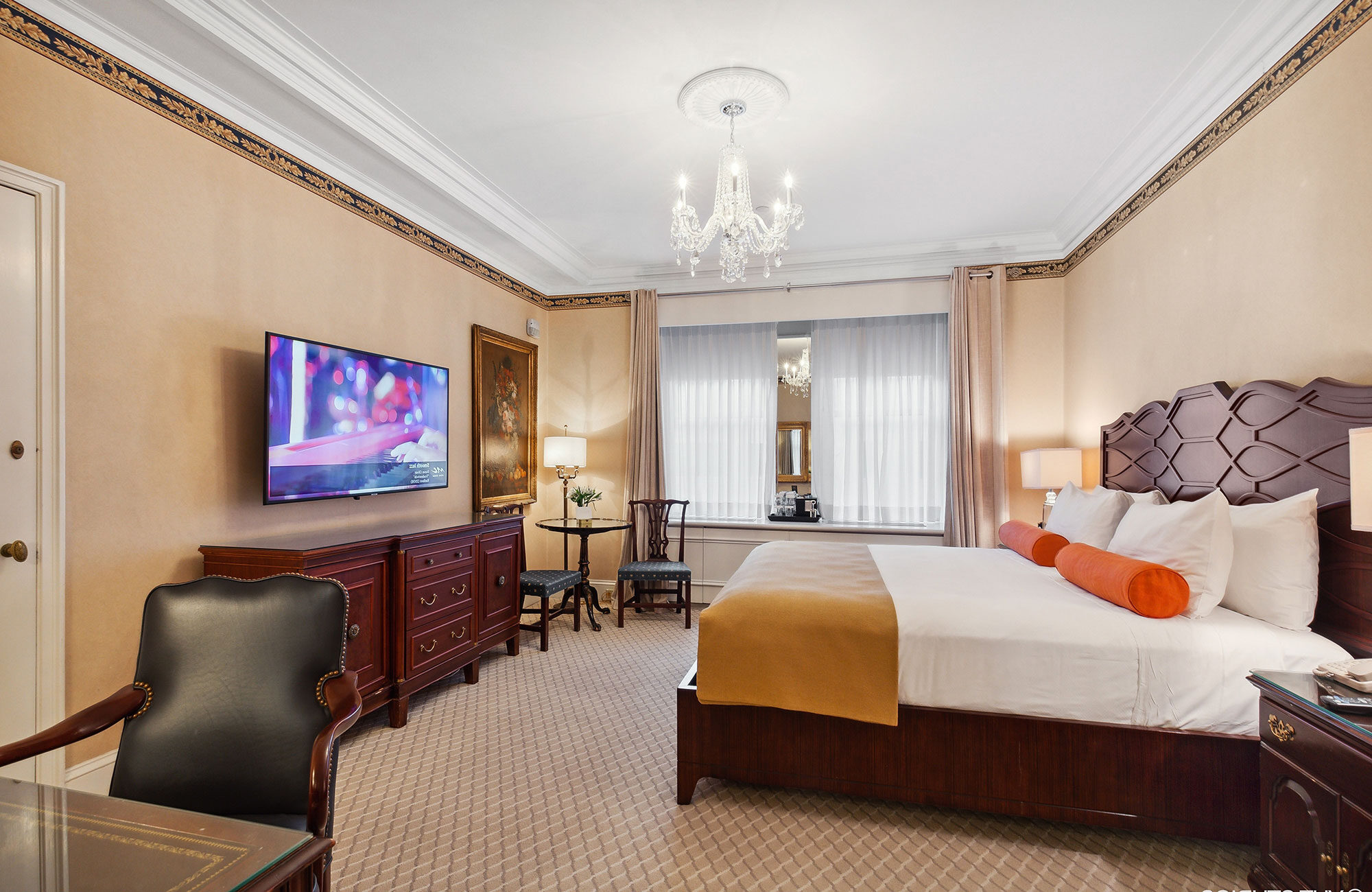 Traditional hotel room with a carved wood headboard, orange bolster pillows, crystal chandelier, and classic furniture including a TV console, writing desk, and small bistro set.