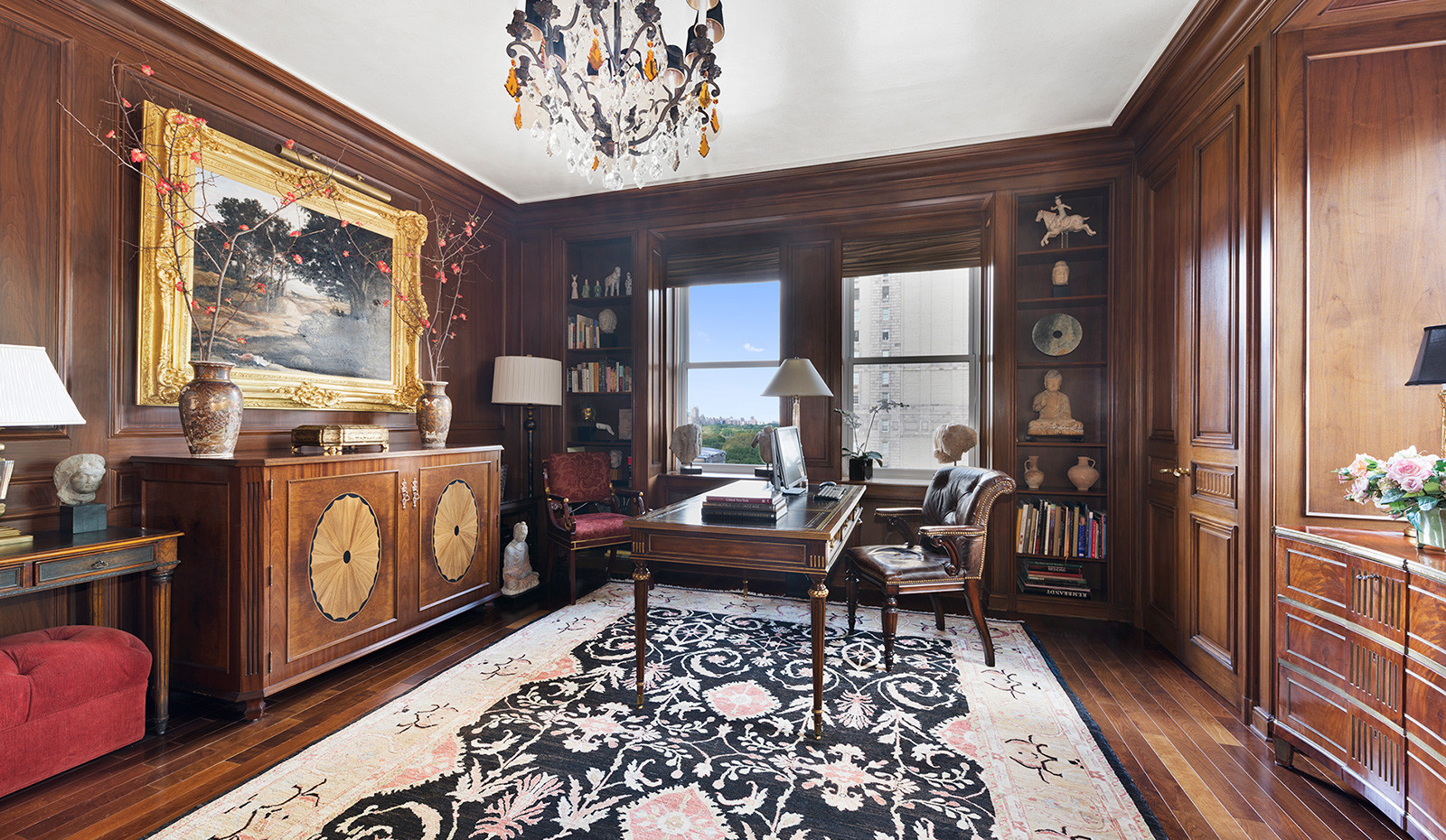 Traditional home office with rich wood paneling, a coffered ceiling, and a chandelier. Features a carved wooden desk, leather chair, and built-in bookshelves framing tall windows with a Central Park view.