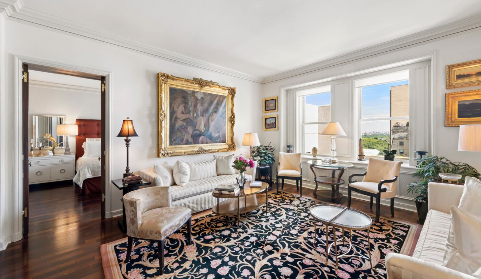 Elegant sitting room with striped cream sofas, a large gilded-framed painting, floral upholstered chairs, a black patterned rug, and large windows overlooking a cityscape. A bedroom with a tufted headboard is visible through an open doorway.