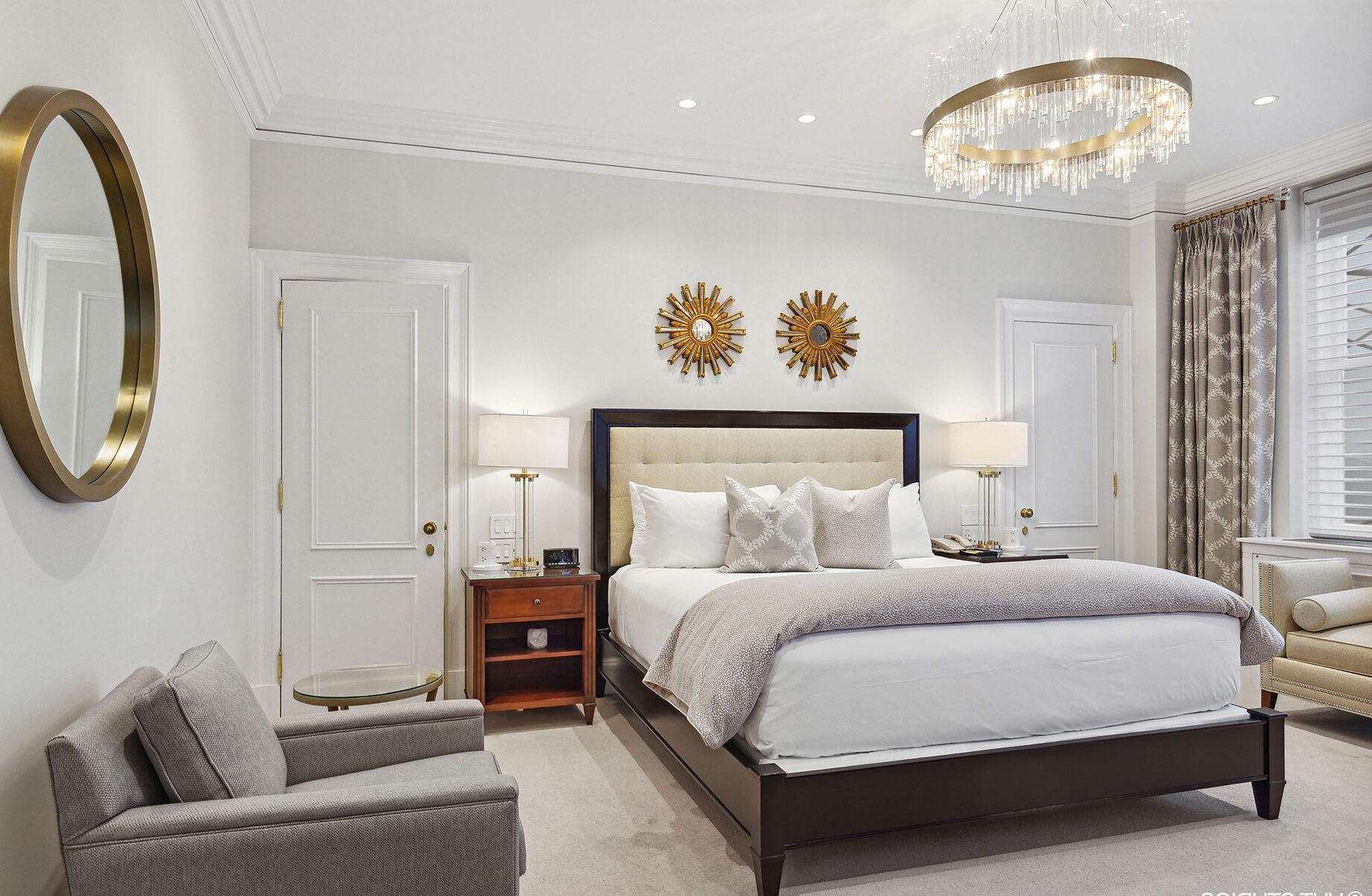 Elegant bedroom with a tufted headboard, sunburst wall decor, neutral tones, crystal chandelier, and plush seating area beside curtained windows.