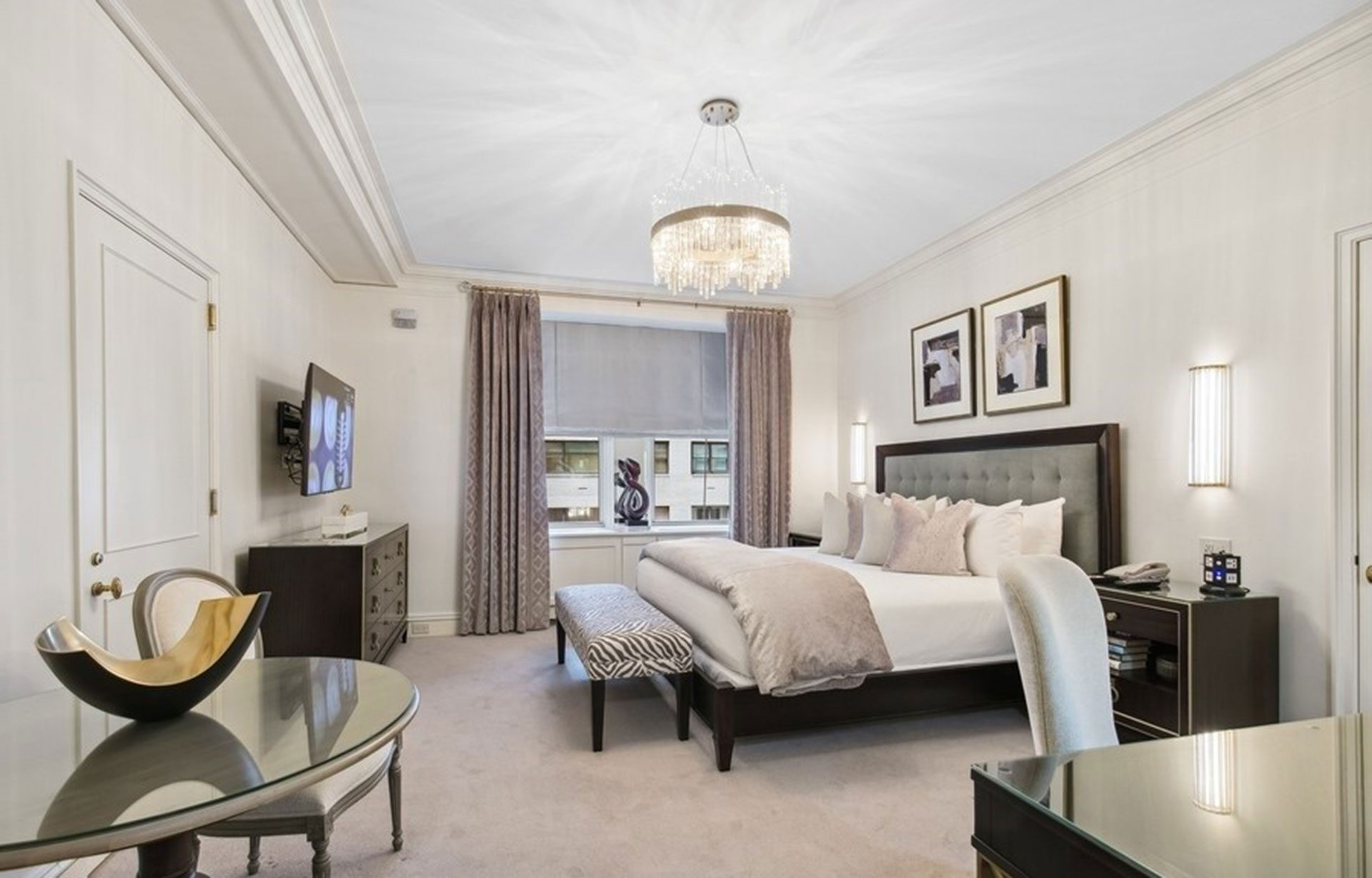 Modern-classic hotel room featuring a tufted headboard, crystal chandelier, soft gray and beige tones, and curated decor with Midtown Manhattan views through the window.