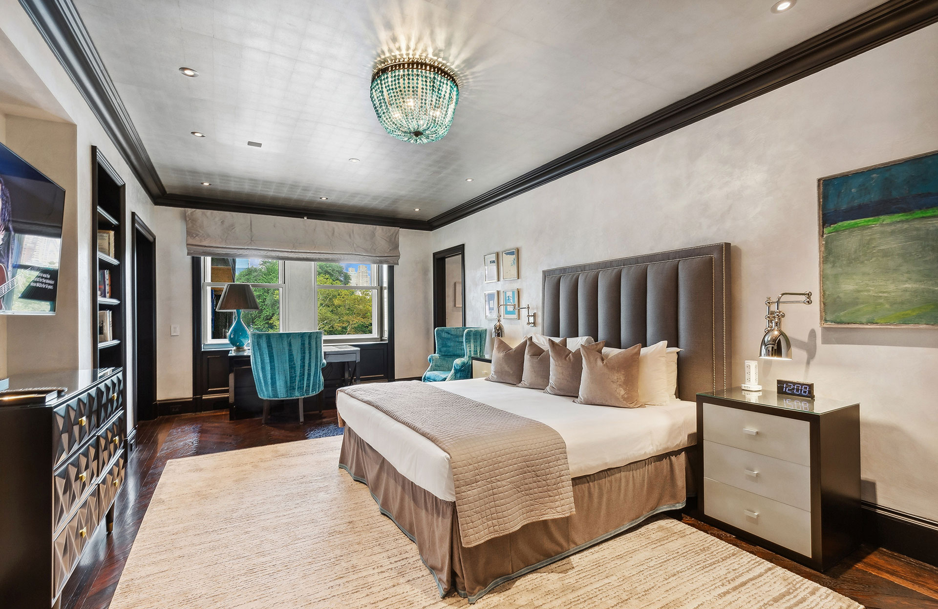 Elegant hotel bedroom with a large upholstered bed, velvet armchairs, a writing desk by the window overlooking trees and skyscrapers, and a modern chandelier casting a soft glow.