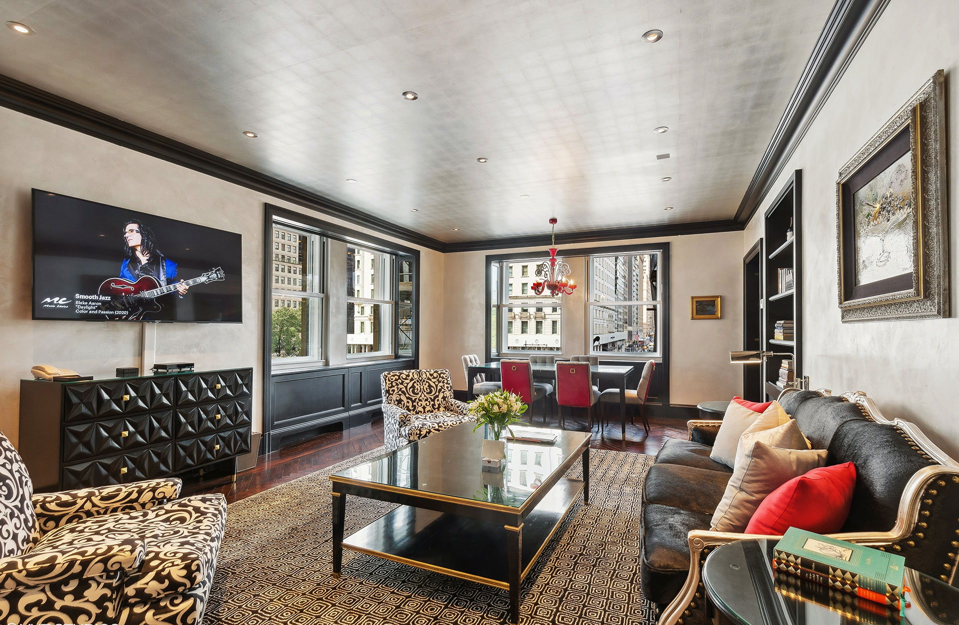 Luxurious hotel living room with black lacquered trim, velvet seating, a glossy black coffee table, a large flat-screen TV, and a dining area framed by oversized windows offering a view of a bustling city street.