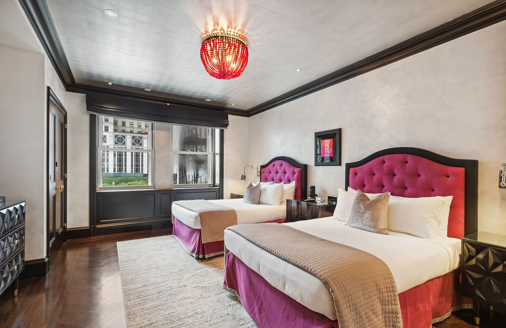 Chic hotel room featuring two double beds with bold magenta tufted headboards, a red beaded chandelier, dark crown molding, and large windows overlooking a city street.