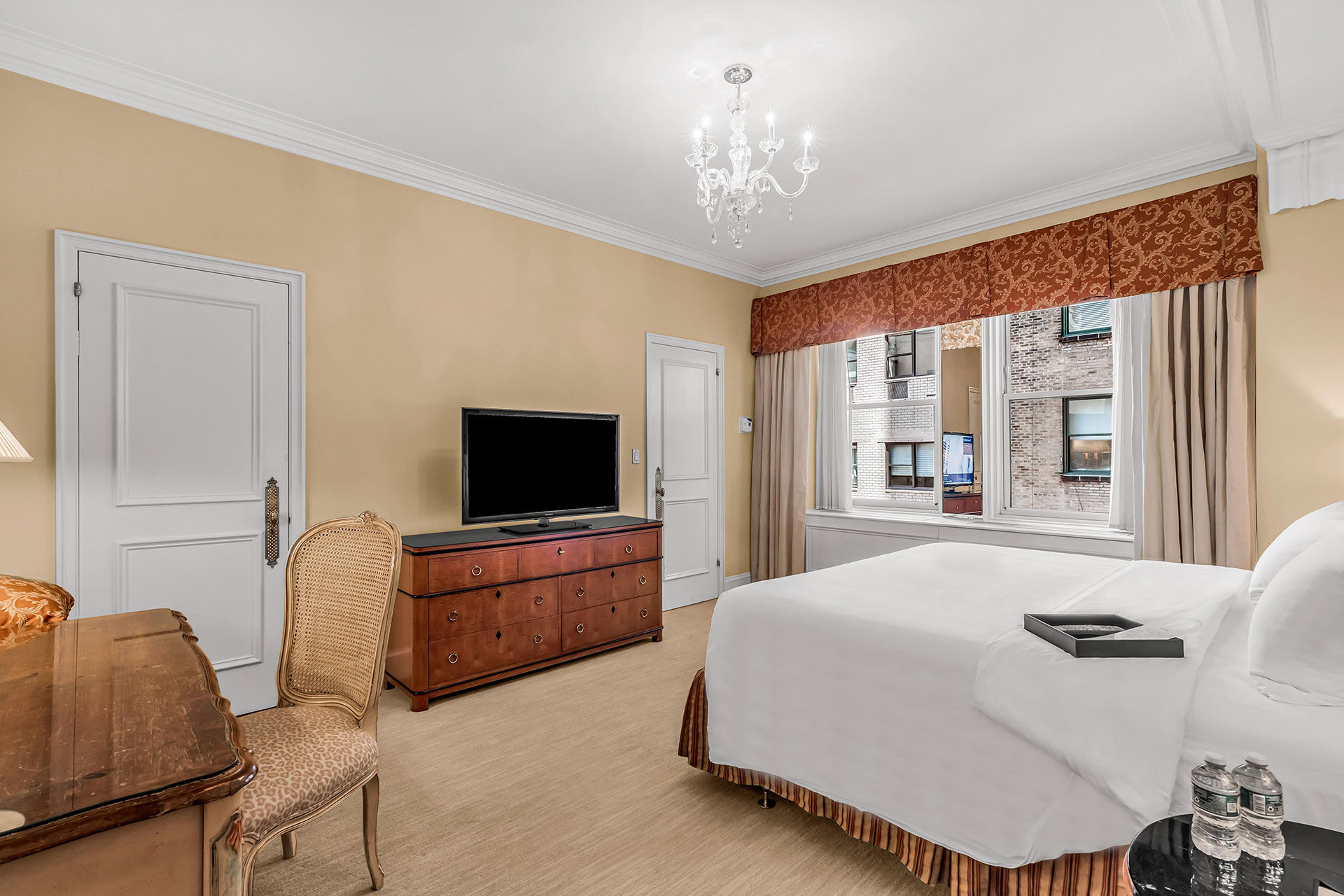 Traditional hotel room with a king bed, wooden writing desk, flat-screen TV on a wooden dresser, and a large window with classic drapery overlooking neighboring buildings.
