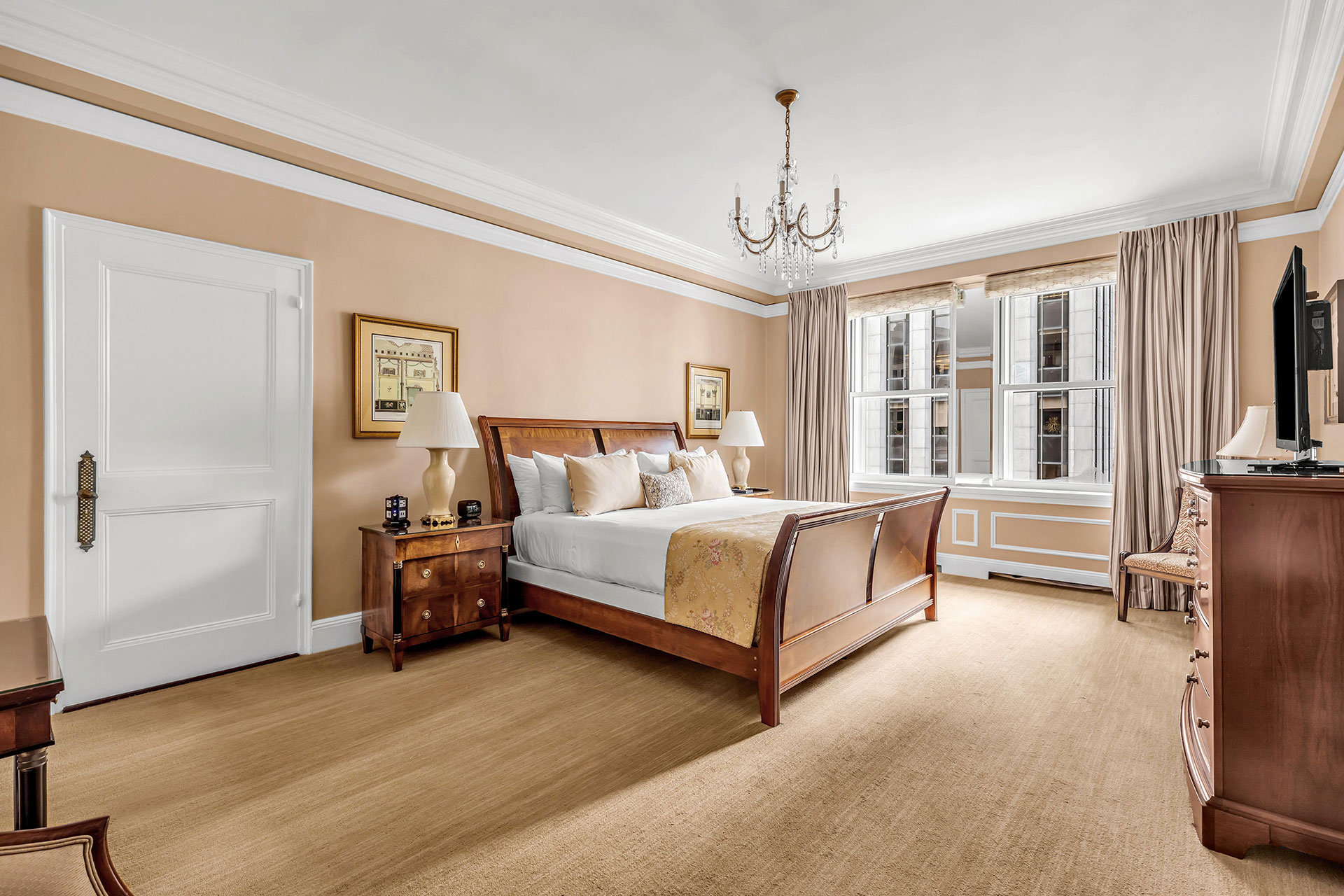 Spacious traditional-style hotel bedroom featuring a sleigh king bed with elegant linens, classic wood furnishings, dual nightstands with lamps, and large windows with floor-length drapes.