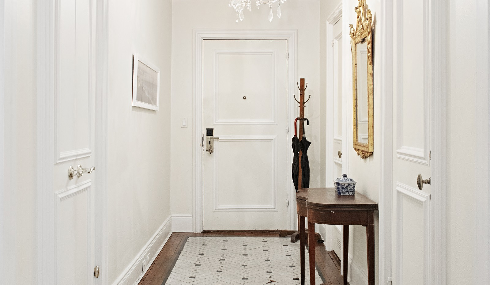 A bright and elegant hallway at The Sherry-Netherland with white walls, dark wood floors, and a patterned rug leading to a white door.