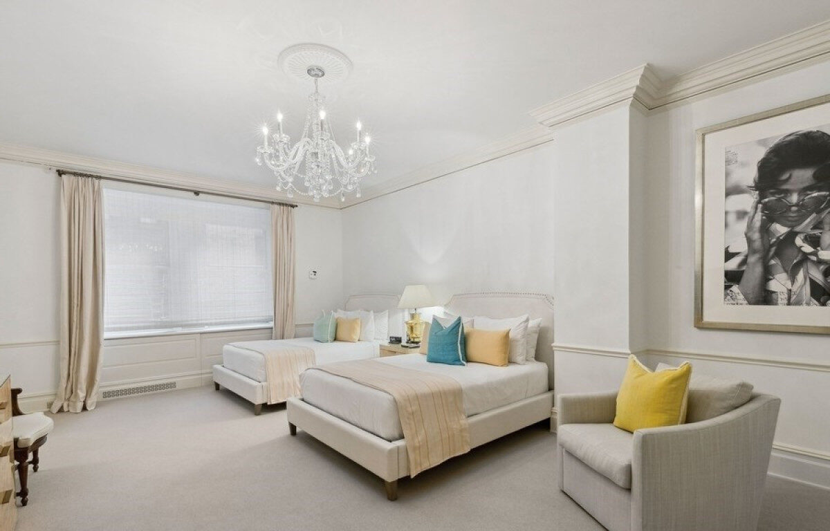 Elegant hotel room with two double beds, soft neutral decor, pastel accent pillows, a crystal chandelier, and a large window with sheer curtains.