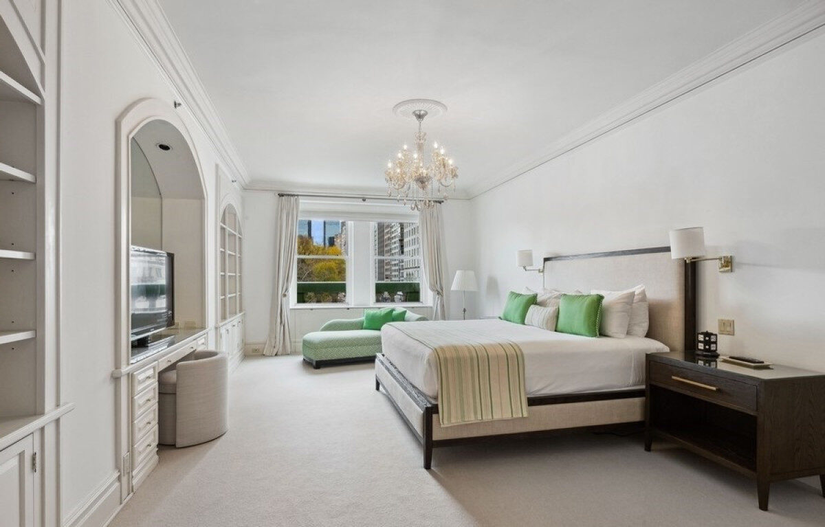 Spacious, sunlit bedroom featuring a king-size bed with green accent pillows, a chandelier, a built-in entertainment unit, and a window offering views of Central Park and city buildings.