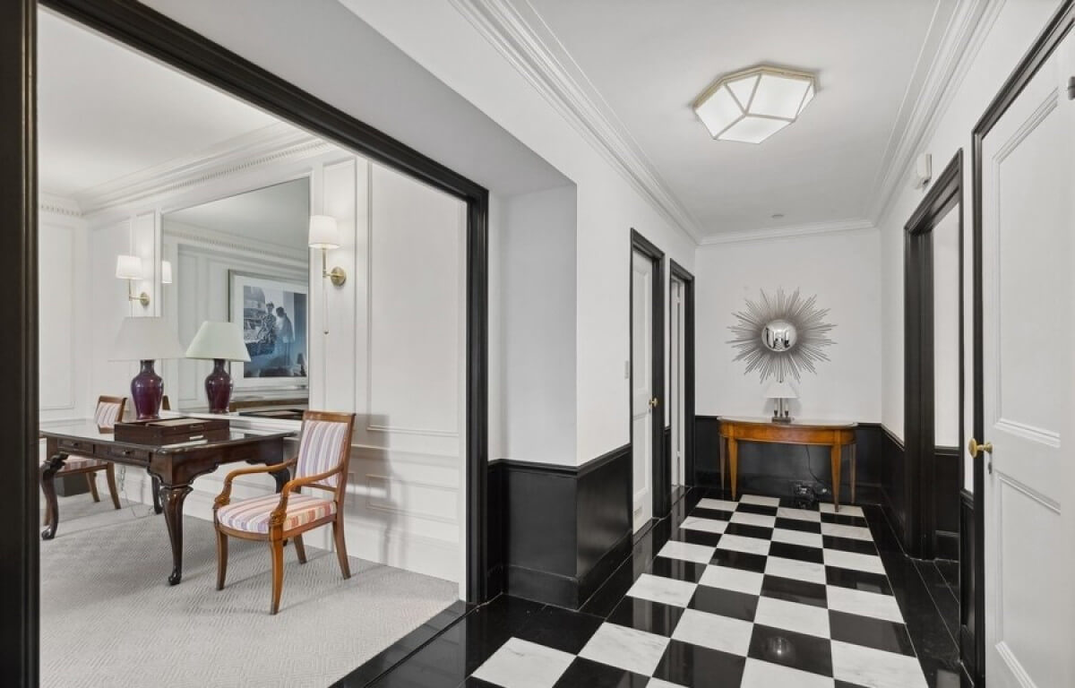 Elegant hotel hallway with black-and-white checkered marble flooring, white walls with black trim, a modern geometric ceiling light, and a mirrored sunburst wall accent above a wooden console table.