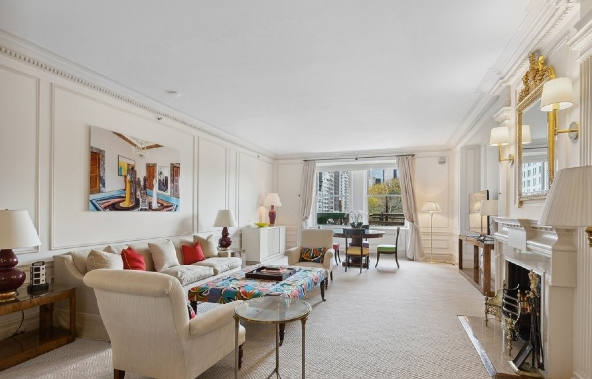 Spacious hotel suite living room with a plush cream sofa, colorful ottoman, elegant fireplace, modern artwork, and large windows overlooking city buildings and Central Park foliage.