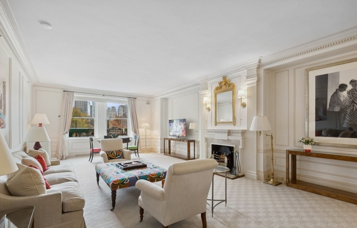 Elegant hotel suite living room with cream armchairs, a colorful patterned ottoman, classic white paneling, a gilded mirror above a fireplace, and large windows revealing Central Park and Midtown Manhattan skyline.